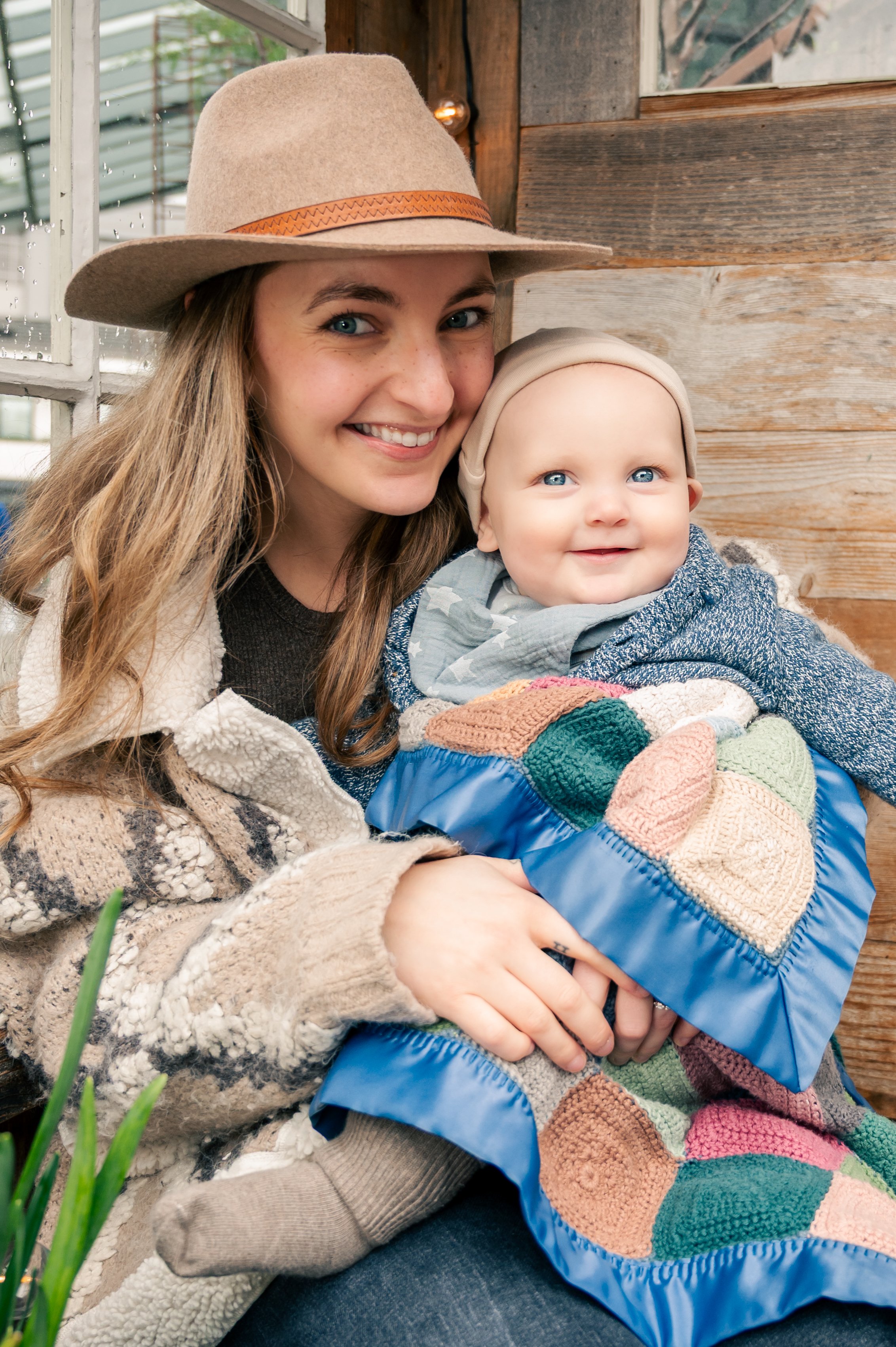 A young woman smiling and holding a baby in a cozy, rustic setting with wooden walls and green plants nearby.