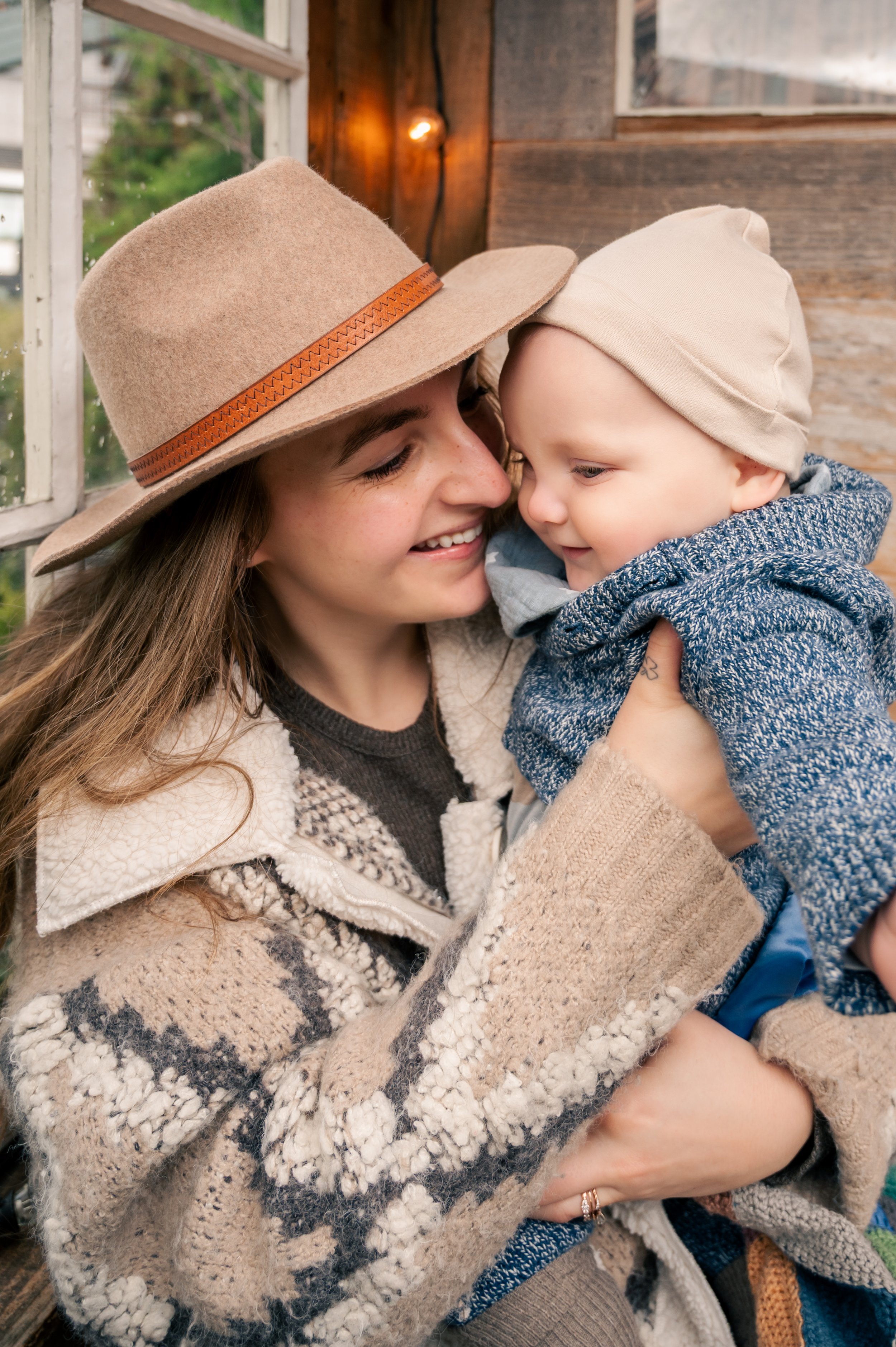 A woman wearing a wide-brimmed hat and a cozy patterned jacket holds a young boy who is wearing a beanie and a knit sweater, both joyfully smiling and touching noses in a warmly lit, rustic indoor setting.