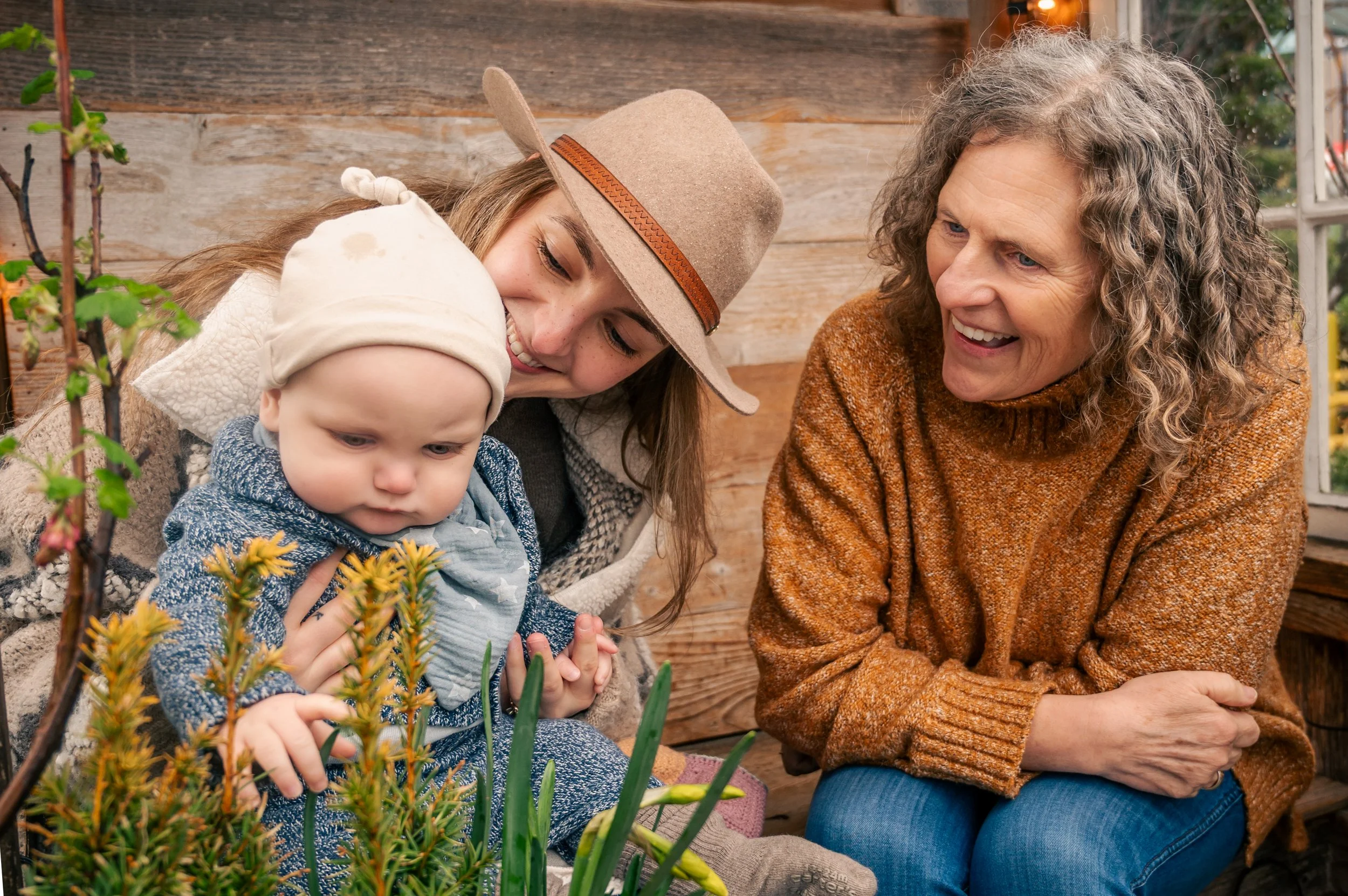 Three generations of women, a grandmother, a young woman, and a baby, spending time together in a garden or greenhouse, surrounded by plants and flowers.