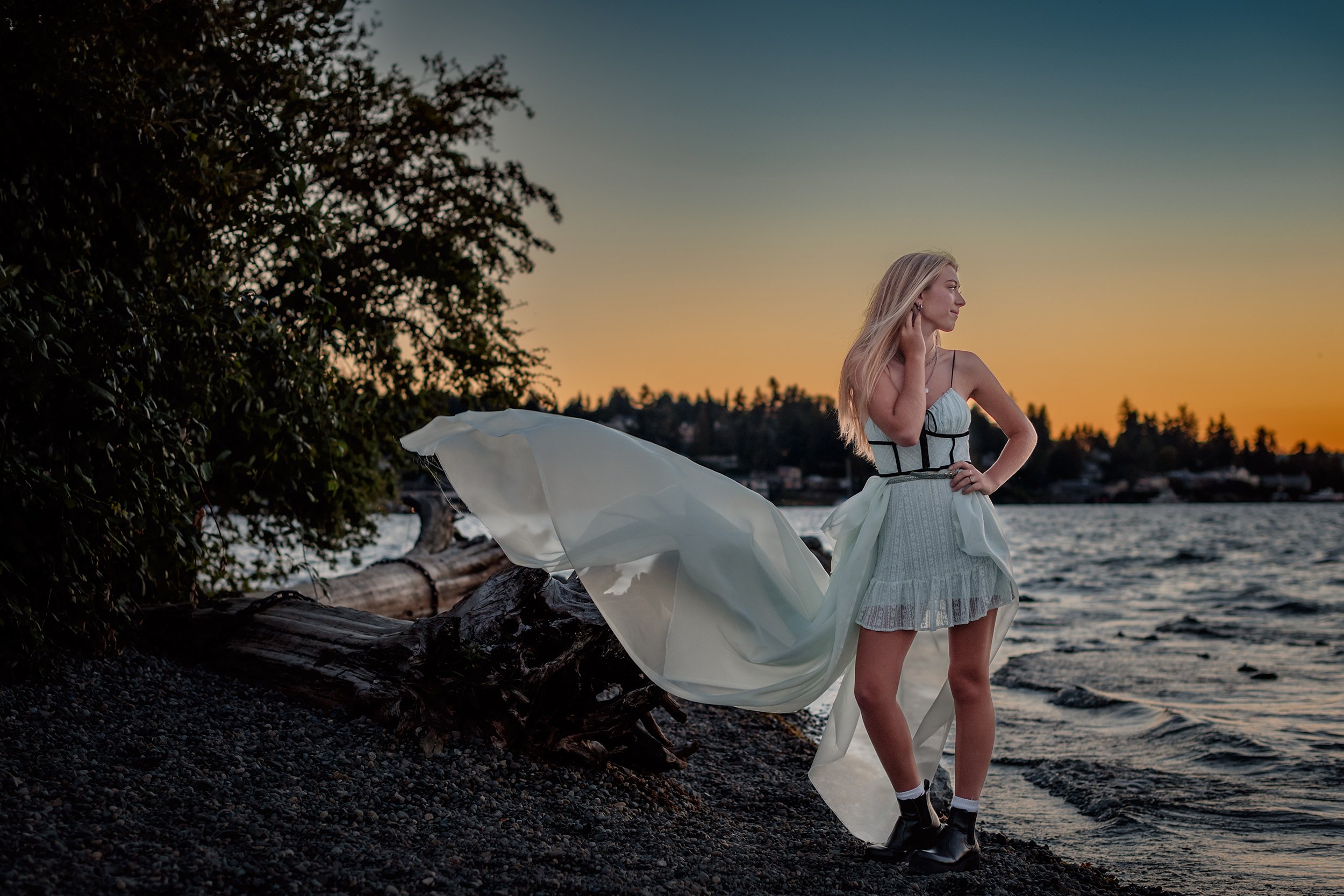 Teenage girl standing on the beach with blue, orange sunset behind her in blue dress with flowing blue train flying in the breeze.