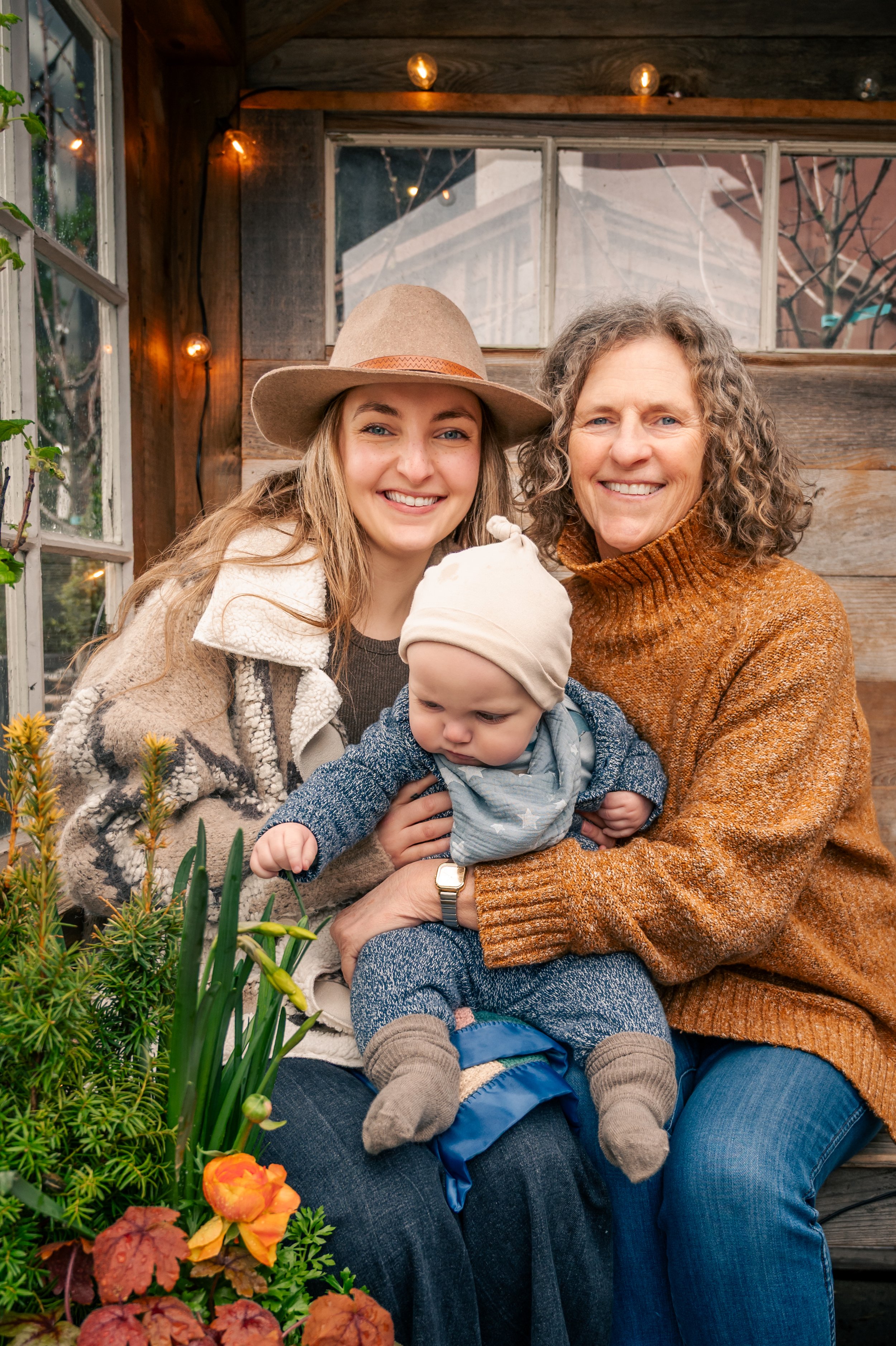 Three generations of a family, a grandmother, mother, and a baby, sitting together on a wooden porch with plants and string lights.