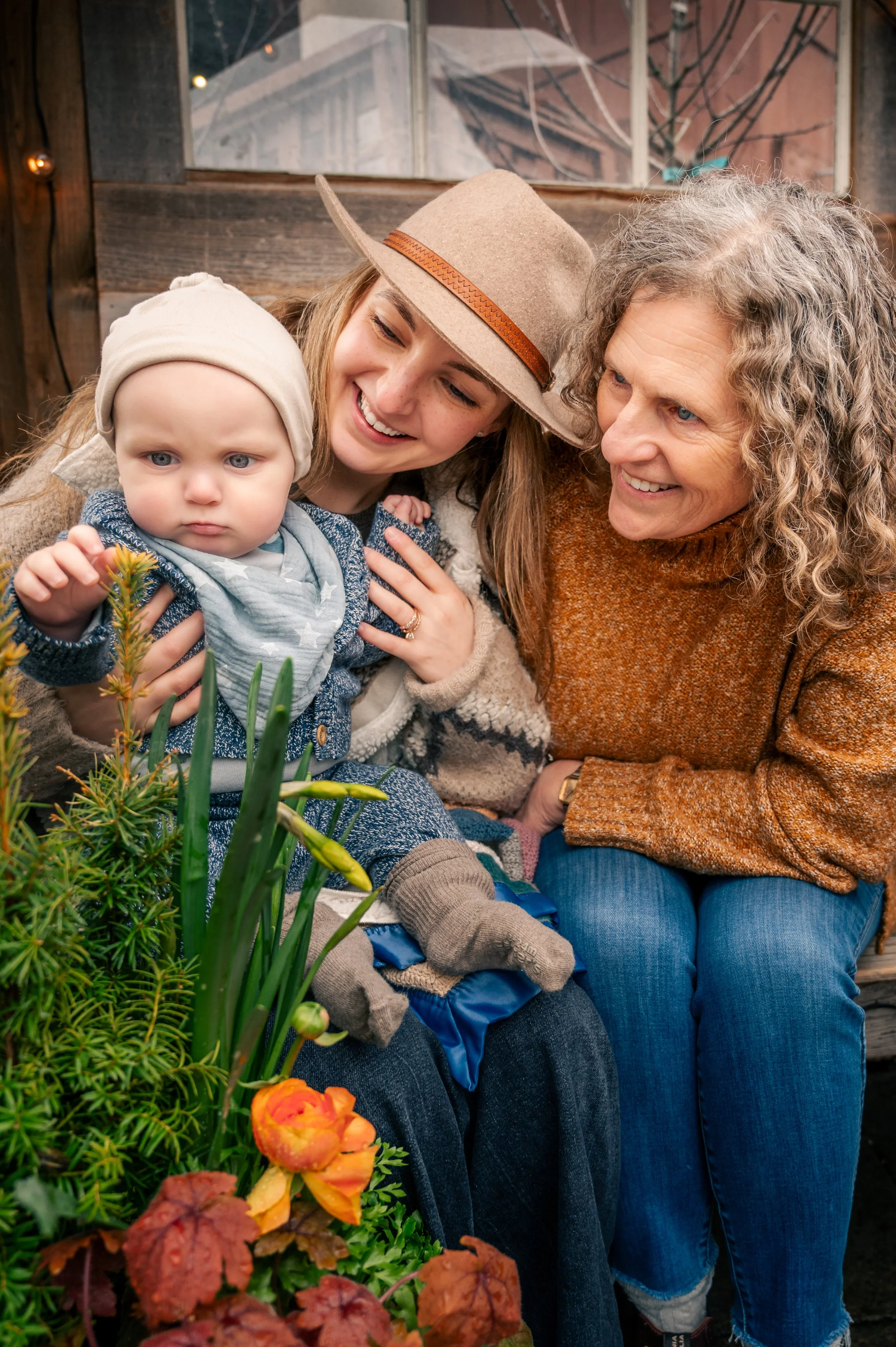 Three generations of women, a baby, a young woman, and an elderly woman, happily gathered outdoors near flowers and greenery, smiling and engaging with each other.