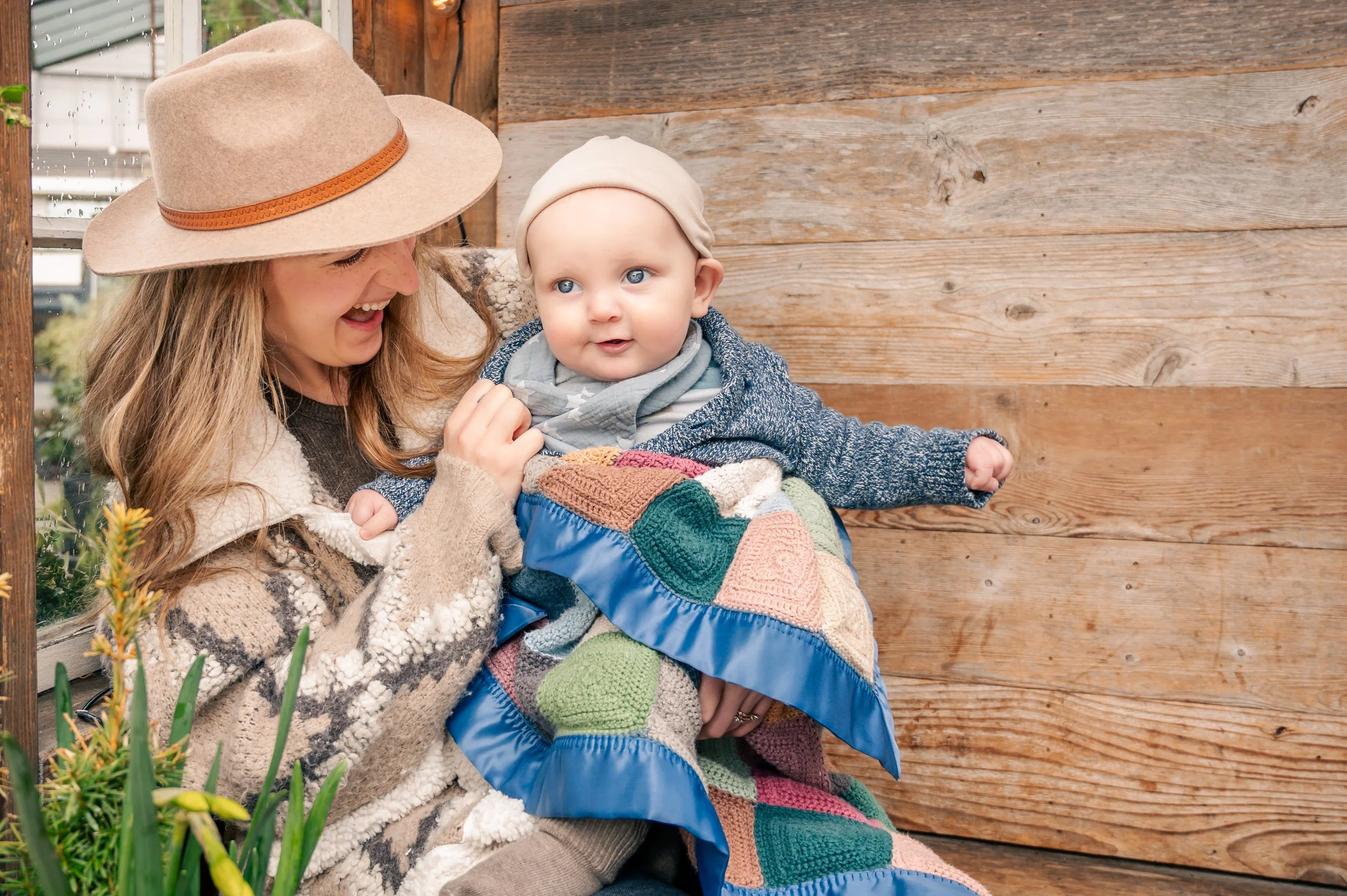 A woman with a wide-brimmed hat holding a baby wrapped in a colorful quilt outside against a wooden wall.