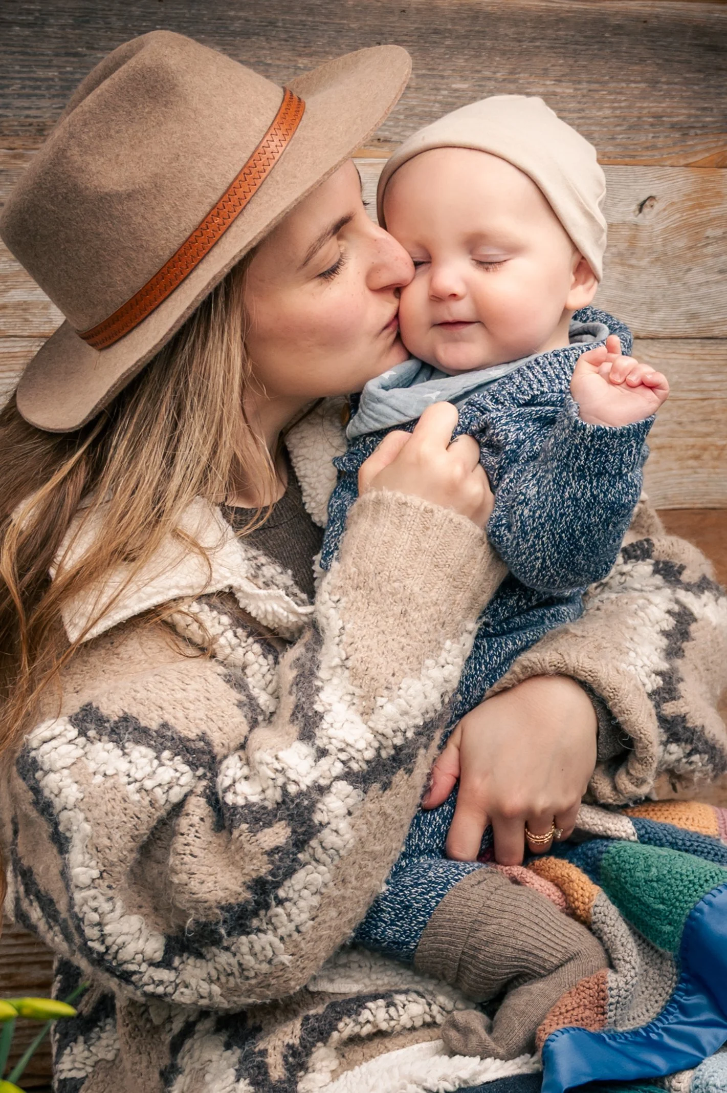 A woman wearing a beige hat and cozy sweater holds a baby, giving a gentle kiss on the nose. The baby, wearing a beige head covering and blue knit sweater, smiles softly with closed eyes, set against a wooden background.