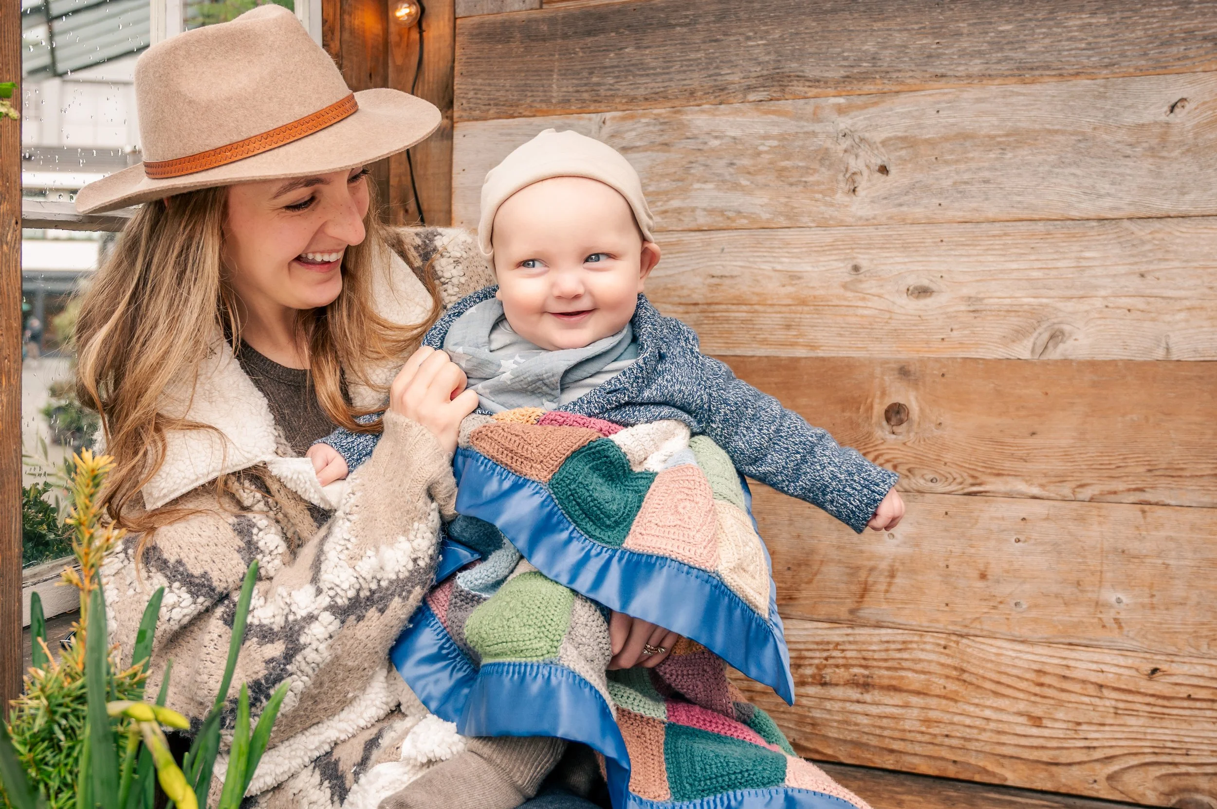 A woman with long hair wearing a beige hat and sweater holds a smiling baby wrapped in a colorful patchwork blanket, sitting outdoors next to a wooden wall.