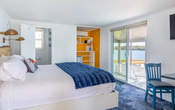 Bedroom with white walls, a large bed with white bedding and blue throw, yellow accent wall with white shelves, sliding glass door leading to balcony with water view, blue chair, and wall-mounted flat-screen TV.