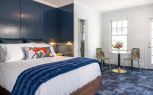 A bedroom with a large bed, blue headboard, and white bedding, including pillows and a blue throw blanket. There is a bedside lamp, and a small table with two chairs near a window with floral arrangement.