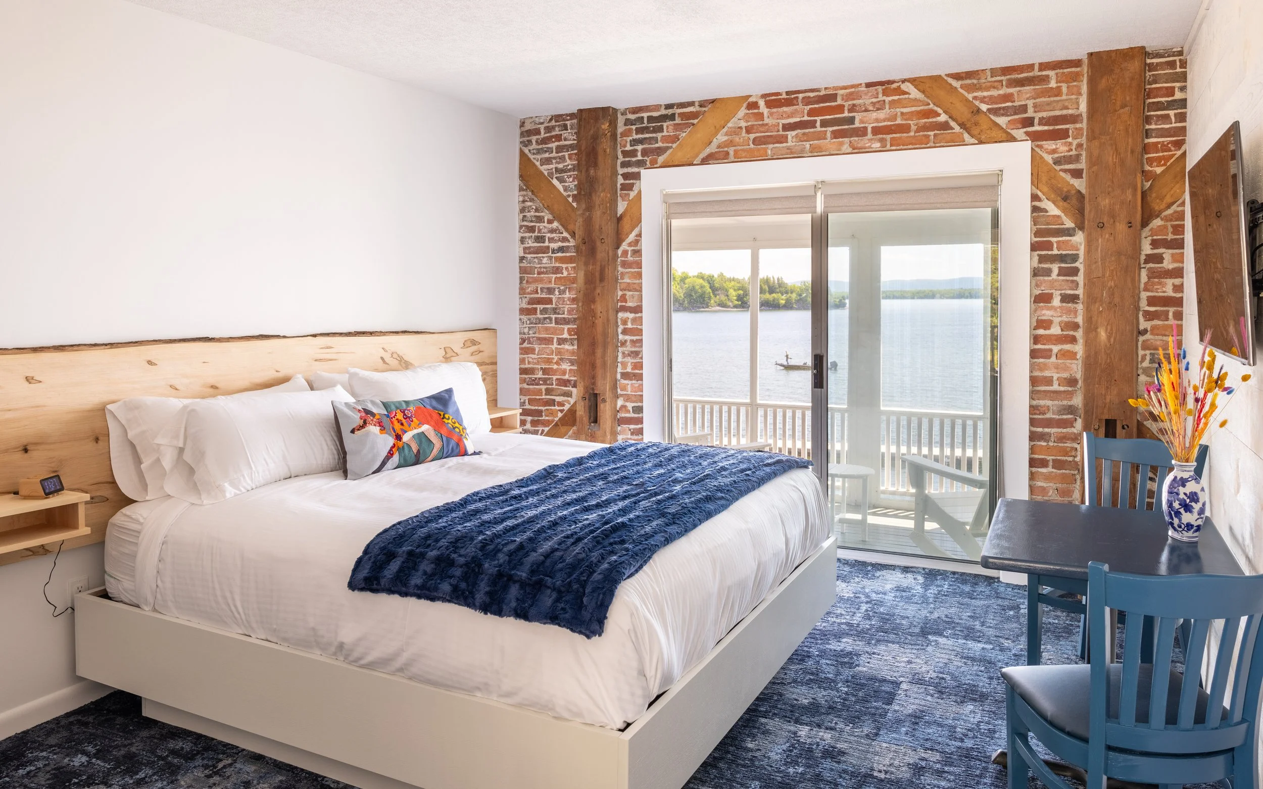 A bedroom with a large bed featuring white bedding and colorful pillows, wooden headboard, brick wall accents, sliding glass doors leading to a balcony overlooking a lake, and a small table with a blue chair and a vase of yellow flowers.