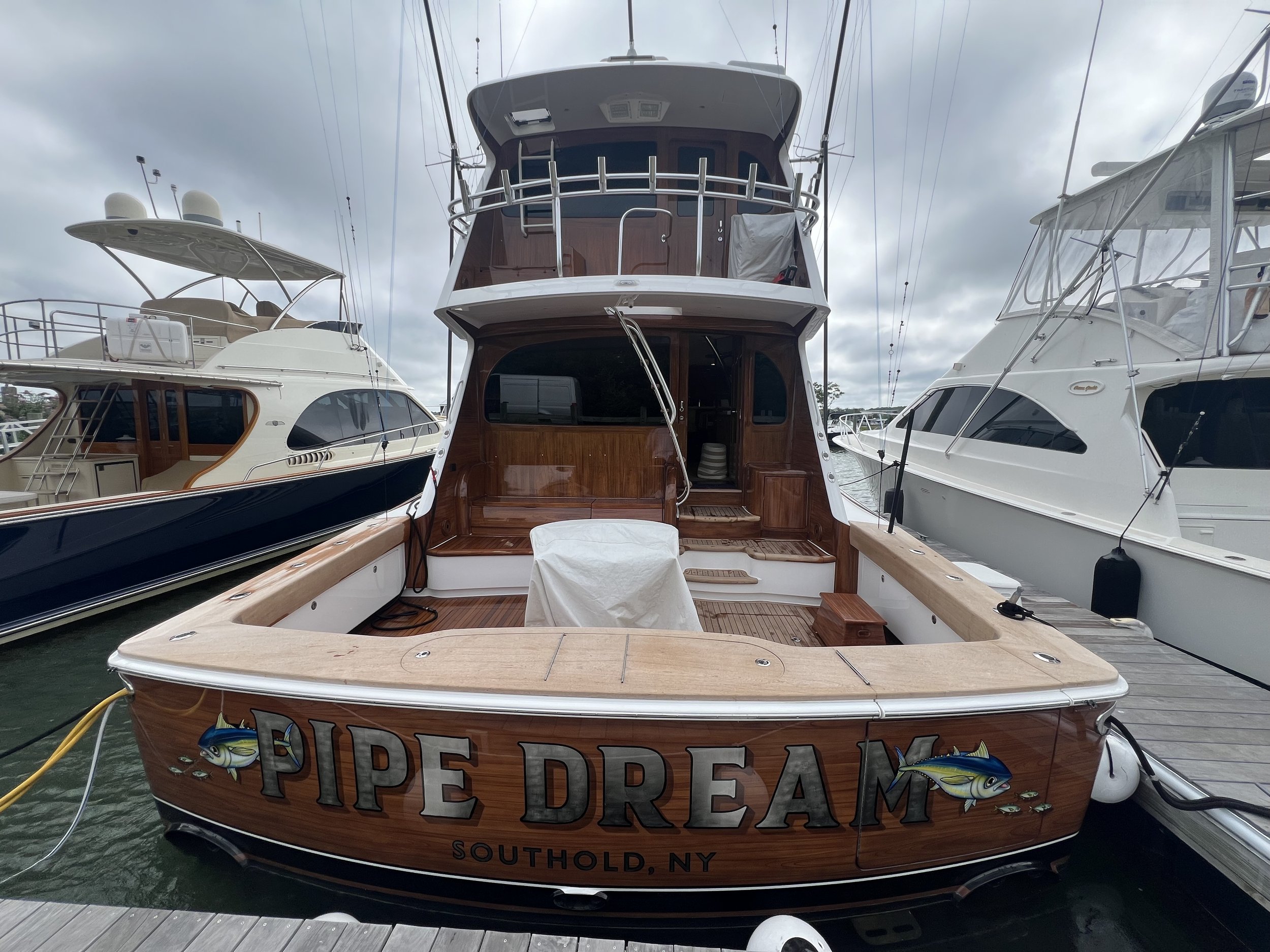 A boat named 'Pipe Dream' docked at a marina, with other boats on either side, under a cloudy sky. The boat is made of wood with a sign indicating it is from Southold, NY.