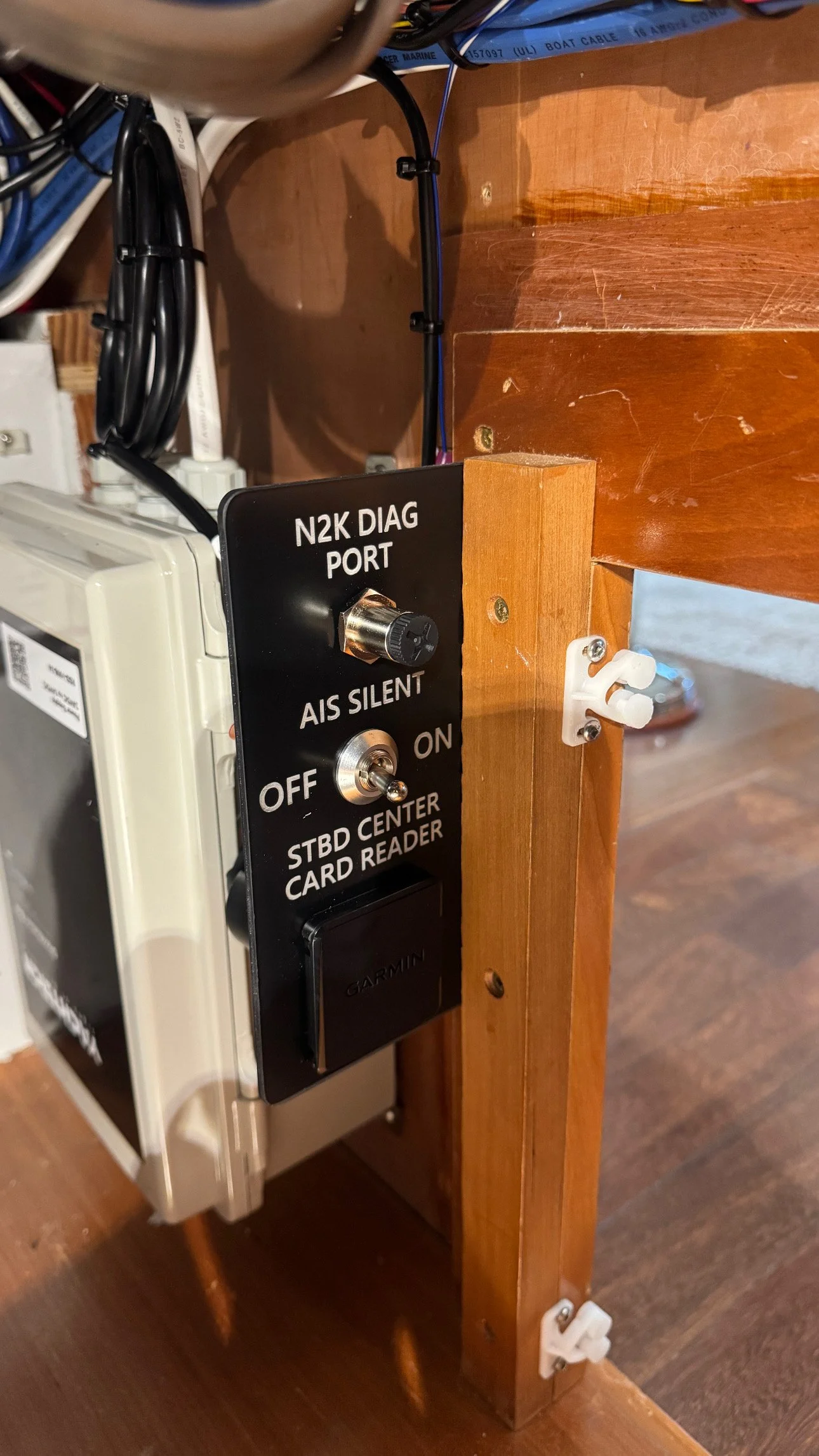 A control panel mounted on a wooden stand, featuring a toggle switch labeled 'OFF' and 'ON,' a rotary switch labeled 'AIS SILENT,' and a black cover labeled 'GARMEN,' with wires connected to electronic equipment behind it.