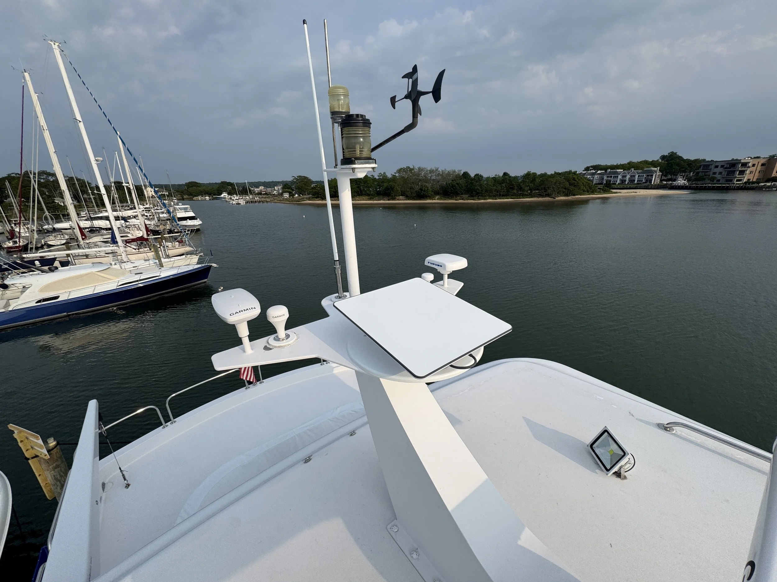 View of a boat's deck with navigation equipment, including GPS, starlink, radar, anemometer, and antennas, docked in a marina with sailboats and houses in the background.