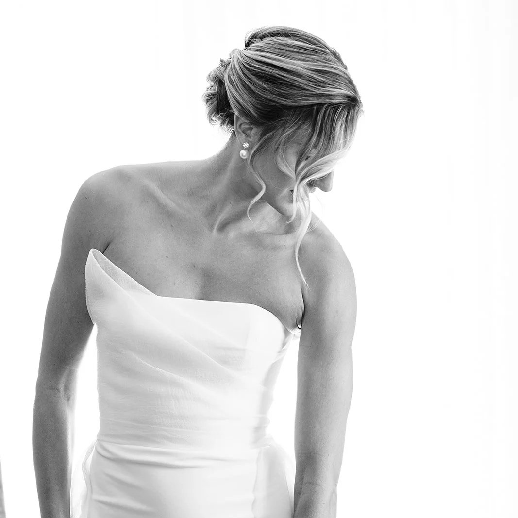 A woman with long brown hair and a cheerful smile standing outdoors in fall attire, wearing a patterned cardigan, white top, and light green pants with her hands in her pockets, with autumn foliage in the background.