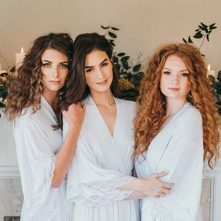 A woman in a white dress with an open back is surrounded by a small group of people outdoors near a wooden fence, with a rustic building and trees in the background, under a clear blue sky.