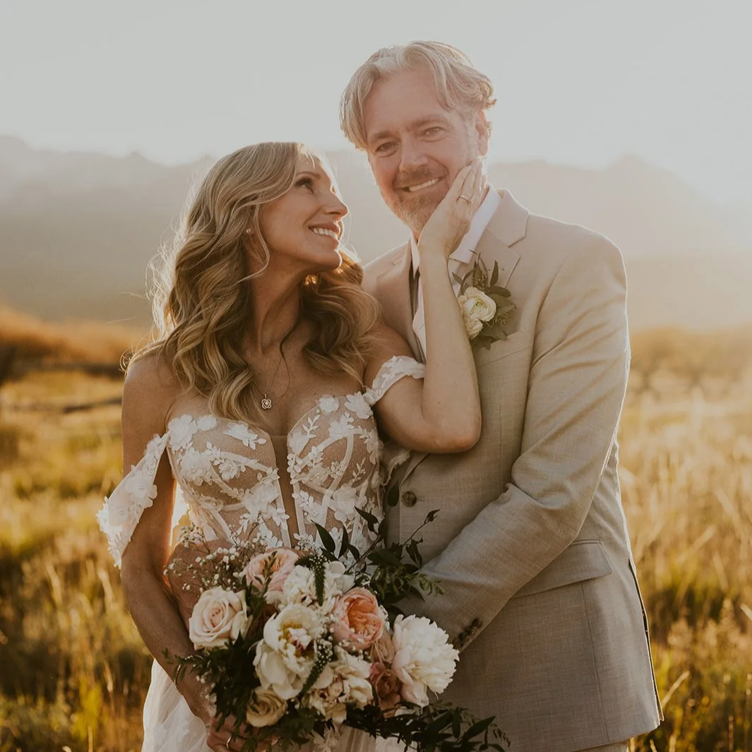 A woman being assisted by a stylist with her hair while another person, possibly a groom, watches as they prepare outdoors on a sunny day.