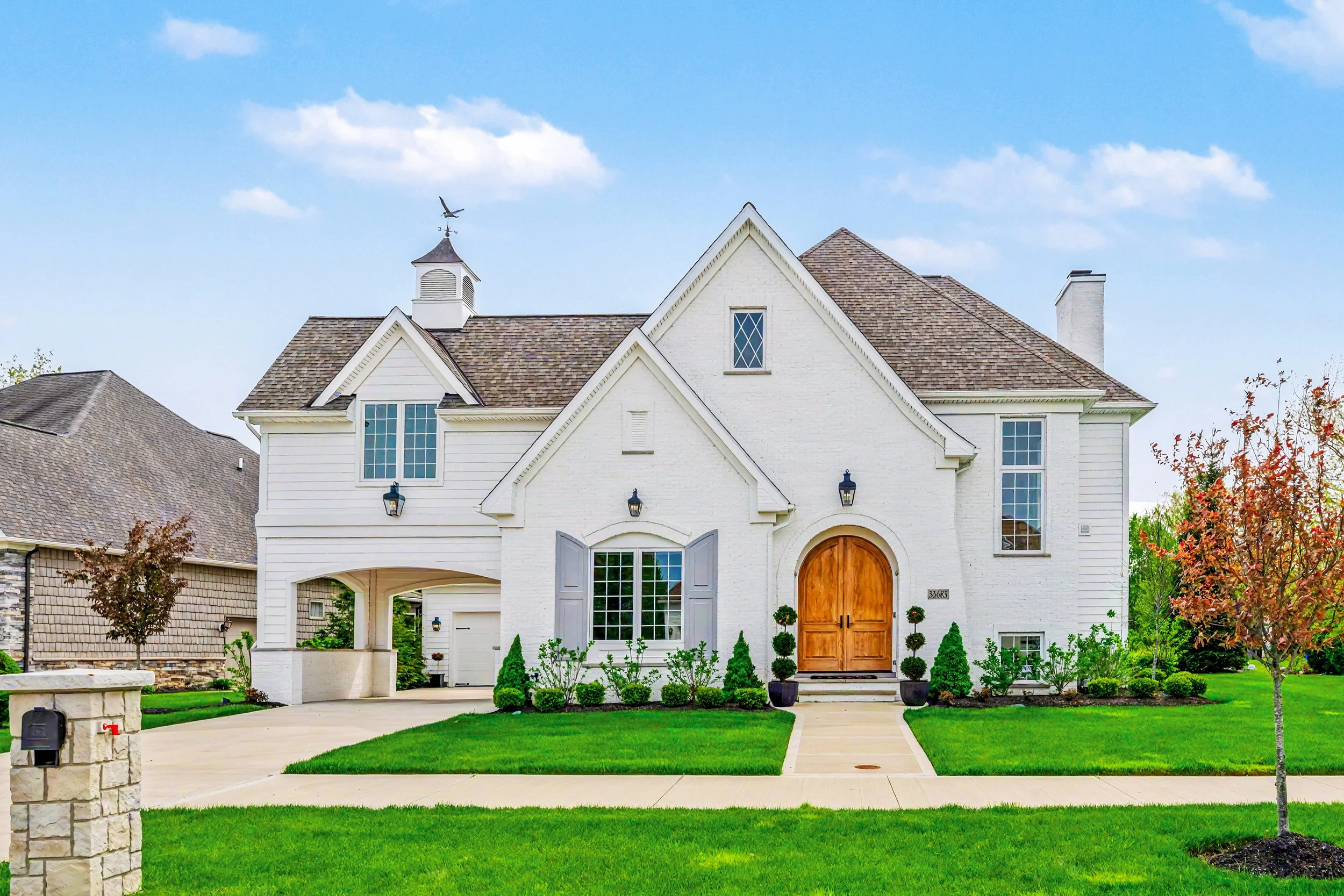 A white, two-story house with a brown shingle roof, arched and rectangular windows, a wooden front door, and a well-manicured lawn with small trees and shrubs.