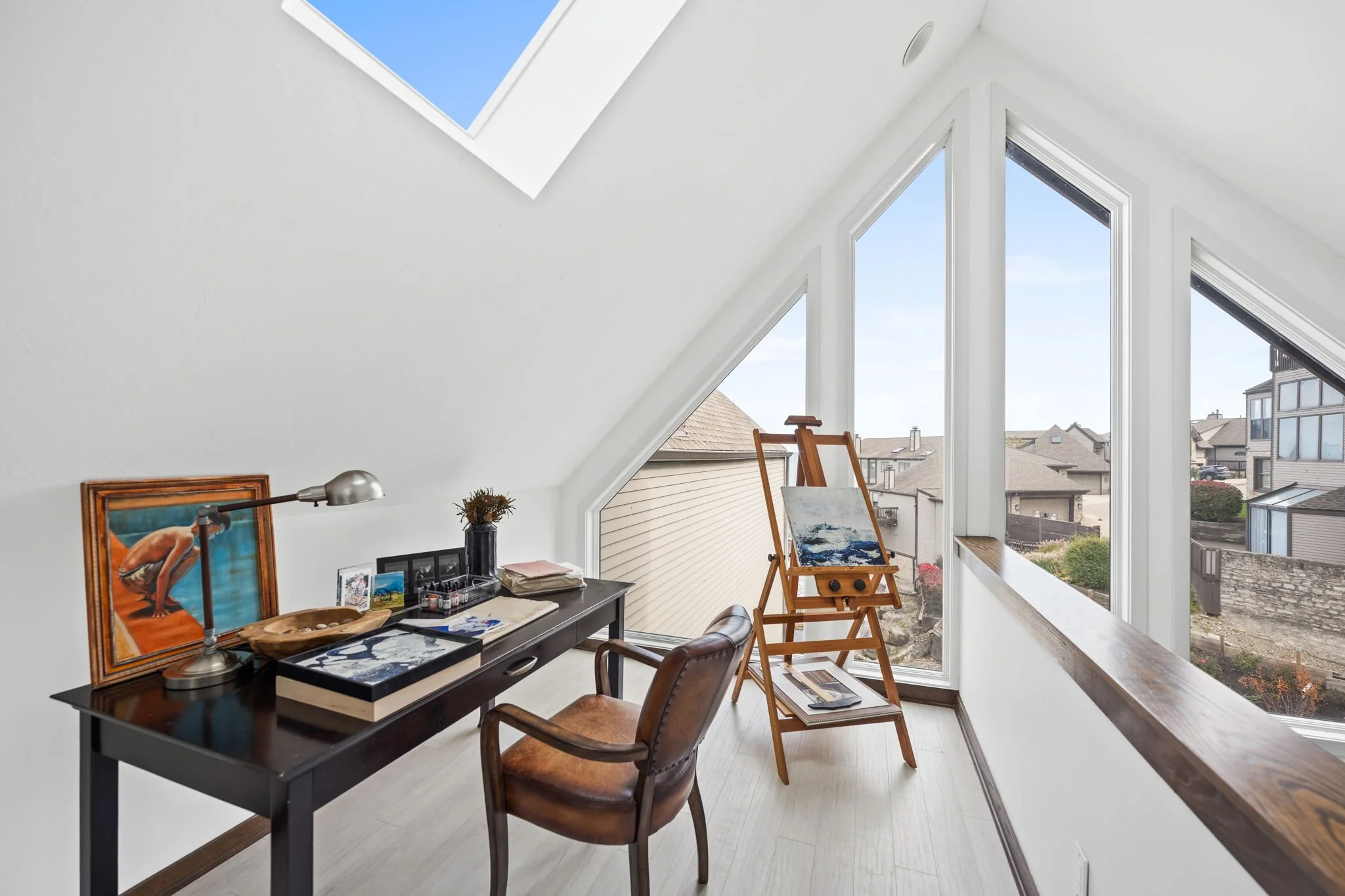 A bright attic room with a sloped ceiling and large windows showing a suburban neighborhood view. The room contains a black table with art supplies, a framed painting, a desk lamp, and a couple of chairs, with an easel near the windows.