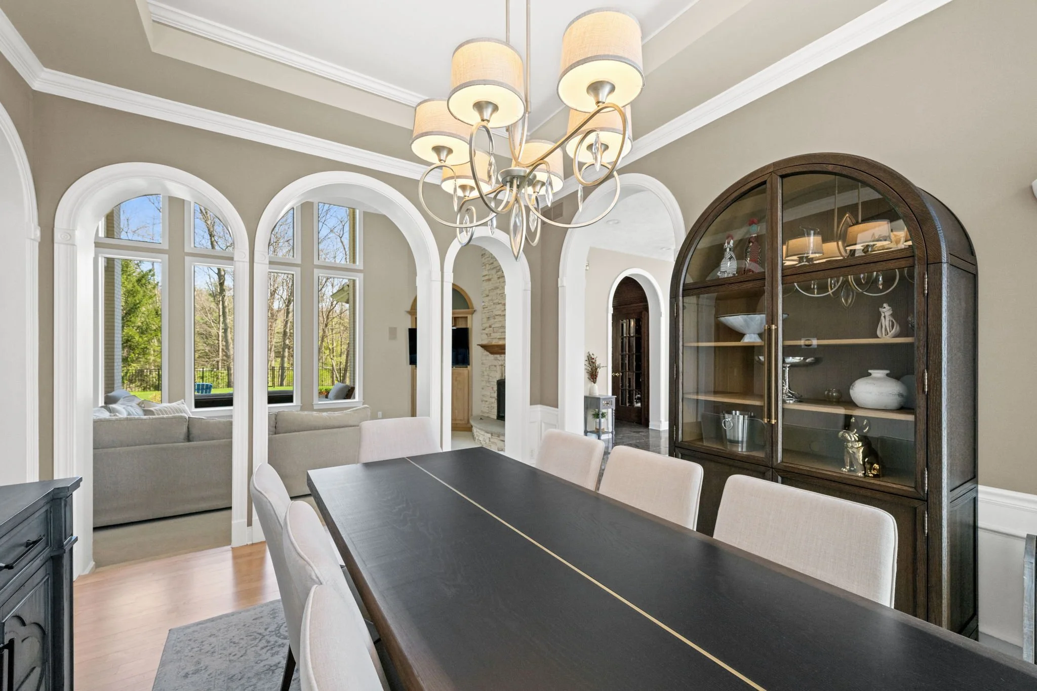 A spacious living room with large arched windows, a modern dining table with white chairs, a black china cabinet, and a chandelier hanging from the ceiling.