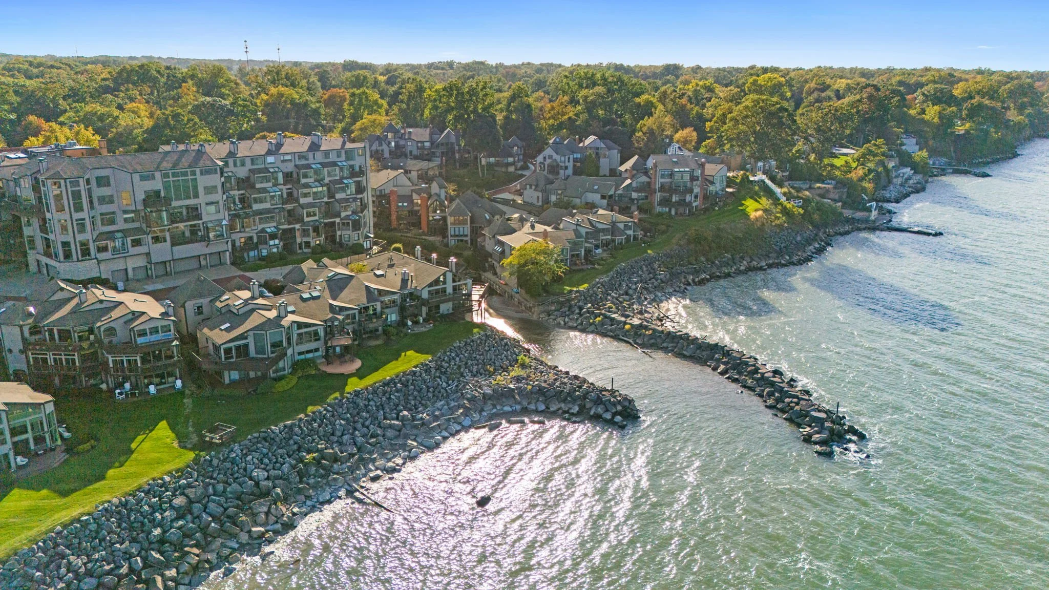 Aerial view of a waterfront residential area with multiple modern houses along the shoreline, a rocky breakwater, and a dense forest in the background on a clear sunny day.