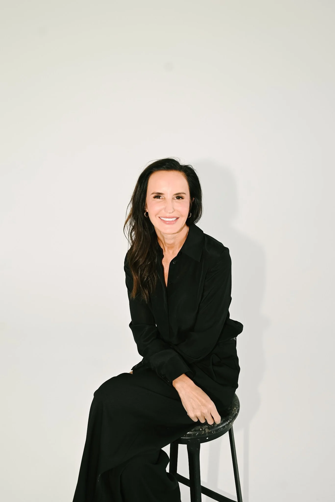 A woman with long dark hair sits on a black stool against a plain white background. She is smiling and wearing a black blouse and pants.