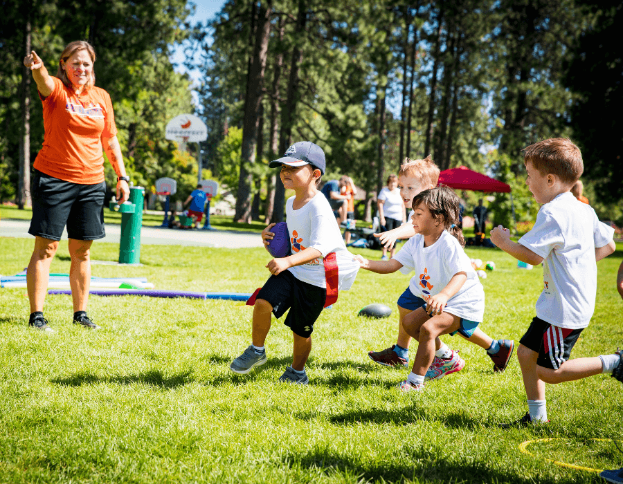 Children participating in a tug-of-war game outdoors on a sunny day, supervised by a woman in an orange shirt.