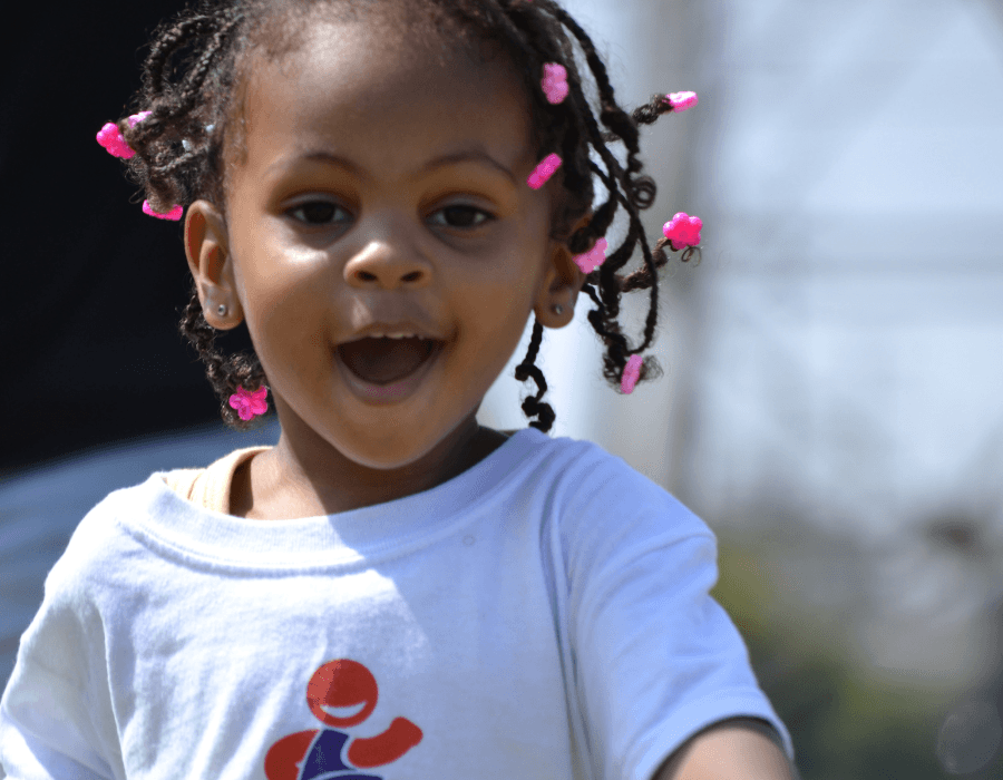 Young girl with braided hair and pink beads, smiling outdoors, wearing a white t-shirt with a cartoon character.