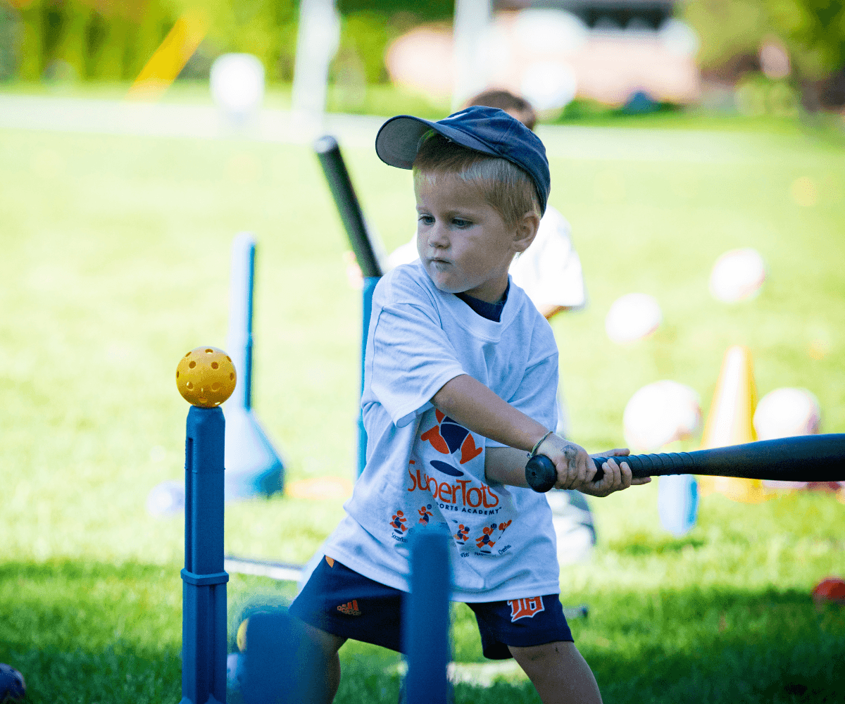 Young boy in a white t-shirt and blue cap swinging a bat at a baseball practice in a park with green grass and colorful cones.