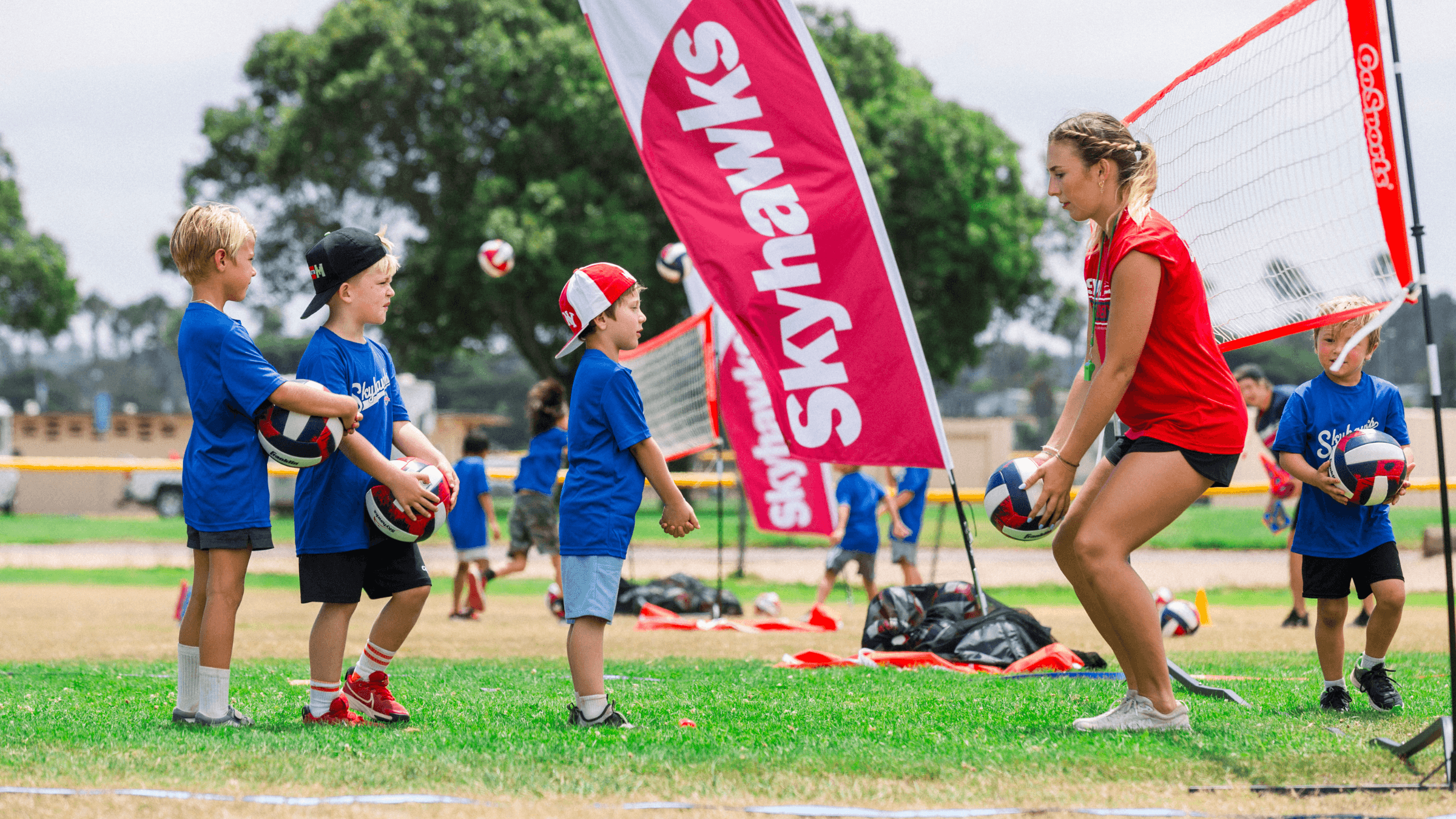 A group of young kids standing in line holding volleyballs during a team practice on a grassy field. An adult female coach in a red shirt and black shorts is preparing a volleyball, with a volleyball net and a pink banner that says 'Skyways' behind her. The scene is outdoors with trees in the background.