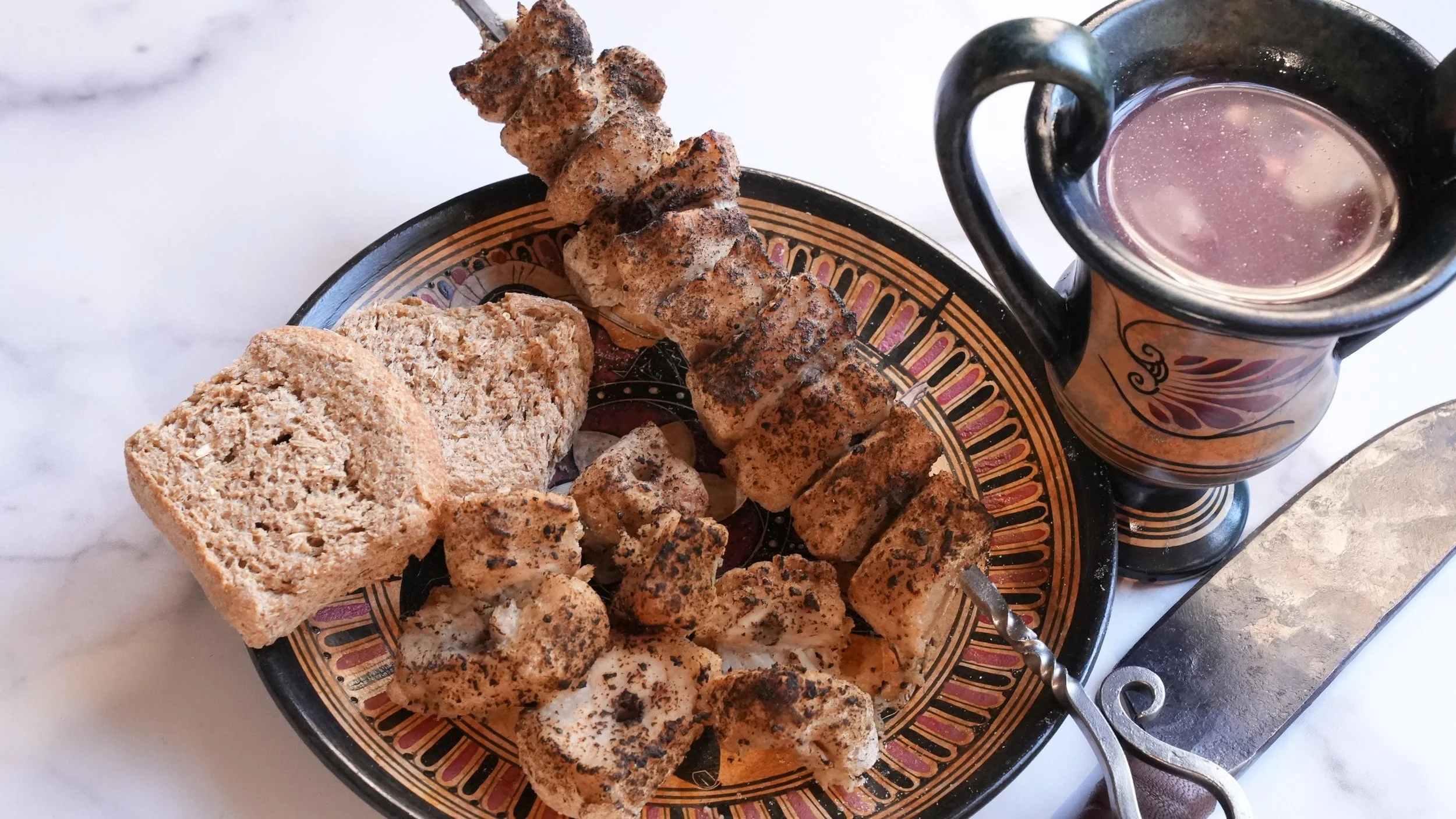 skewer of roasted pork with hard barley bread and a cup of kykeon