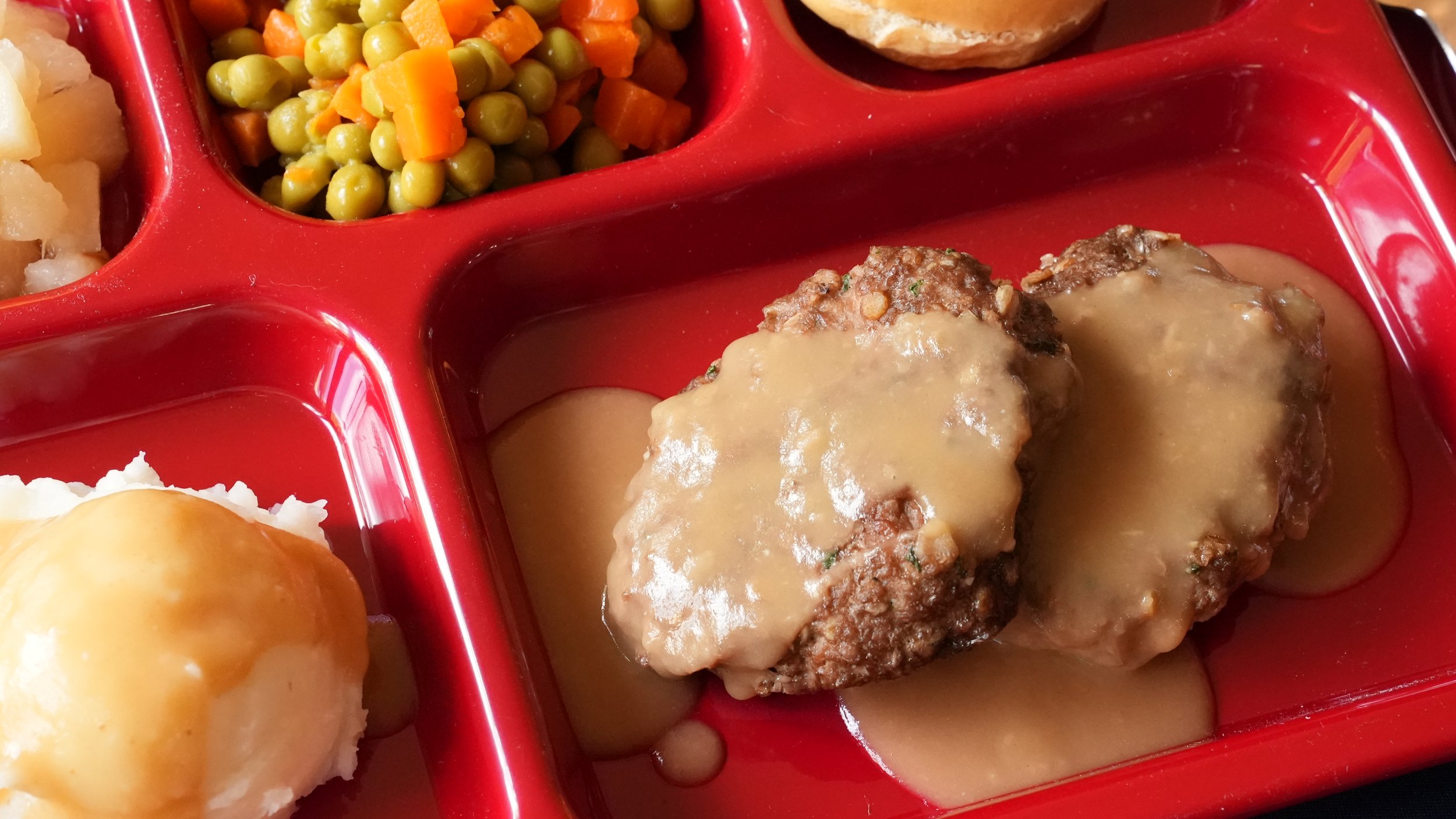 2 salisbury steaks with brown gravy on red lunch tray with mashed potatoes and mixed vegetables