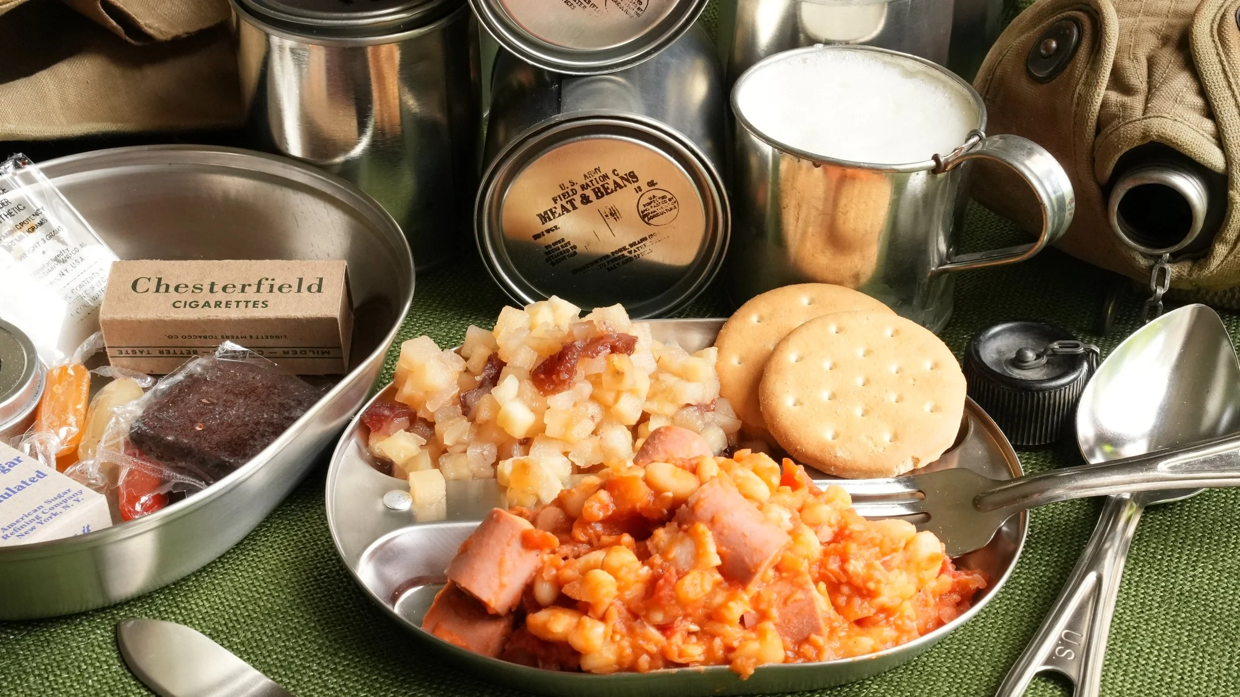 sliced vienna sausages cooked with beans in tomato sauce, apple and cranberry sauce, and 2 crackers in a metal mess kit, and a metal cup of frothy pinapple milk