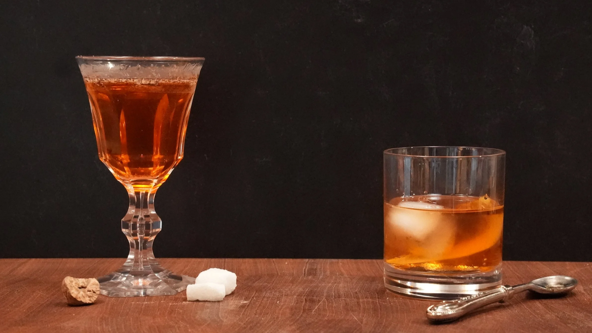 left: amber cocktail in stemmed glass with sugar cubes and nutmeg on the side. Right: amber cocktail in rocks glass with lemon twist ice cube, a silver spoon is on the side