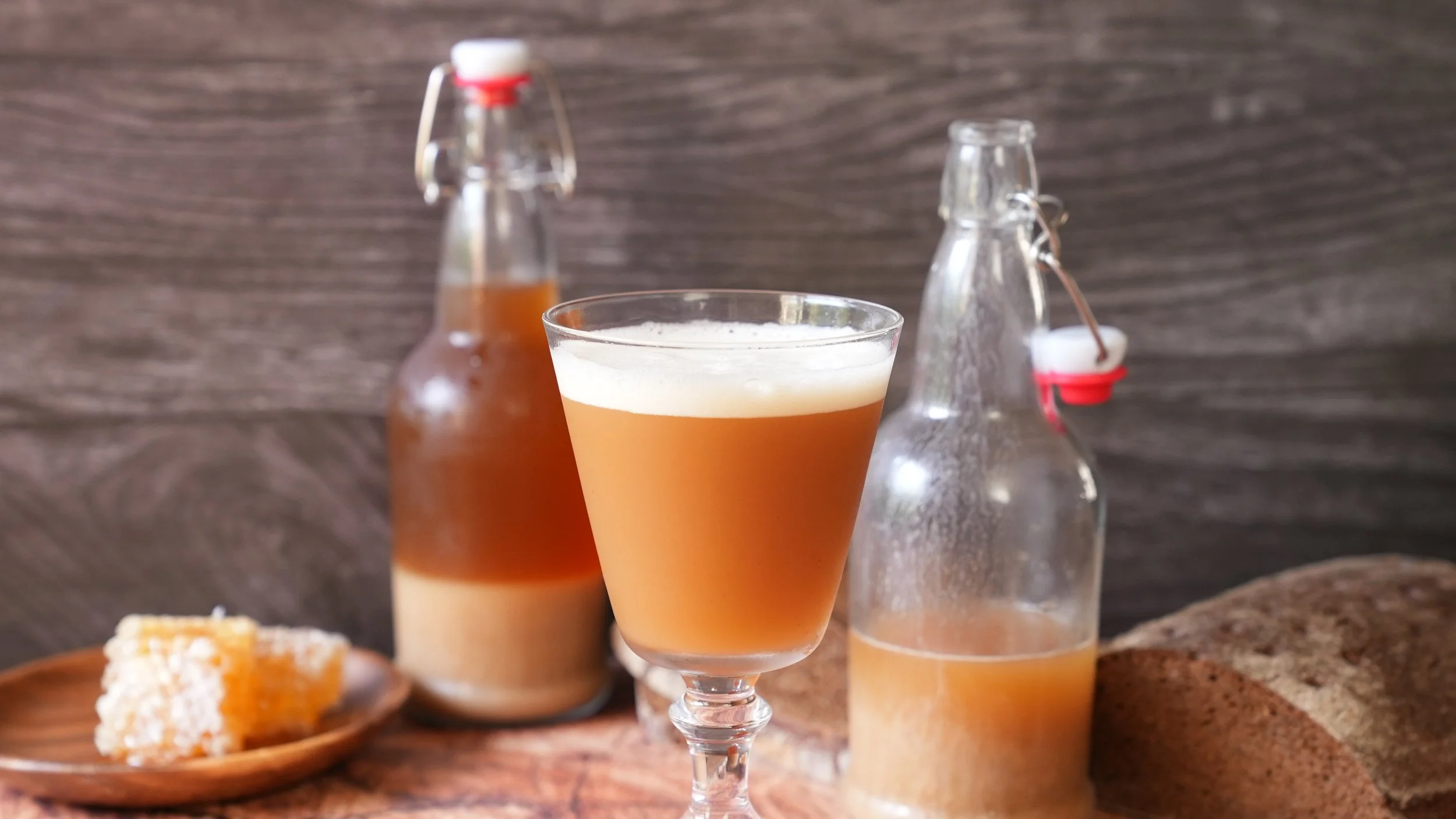 cloudy amber colored kvass with foam on top in a glass with a loaf of dark rye bread, honeycomb, and bottles of kvass in the background