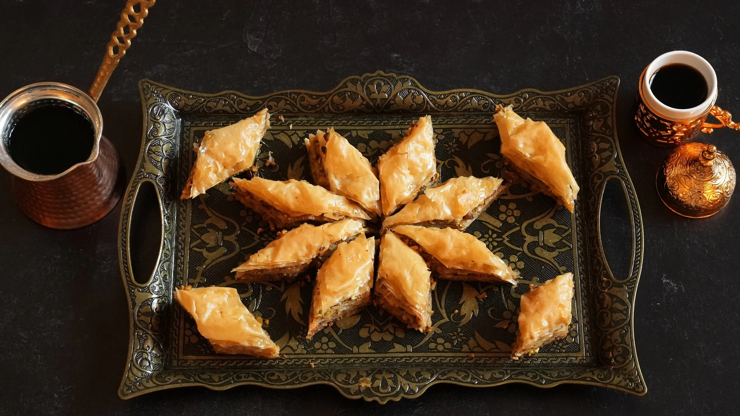 diamond shaped pieces of baklava filled with pistachios on a dark metal tray with a pot of Turkish coffee