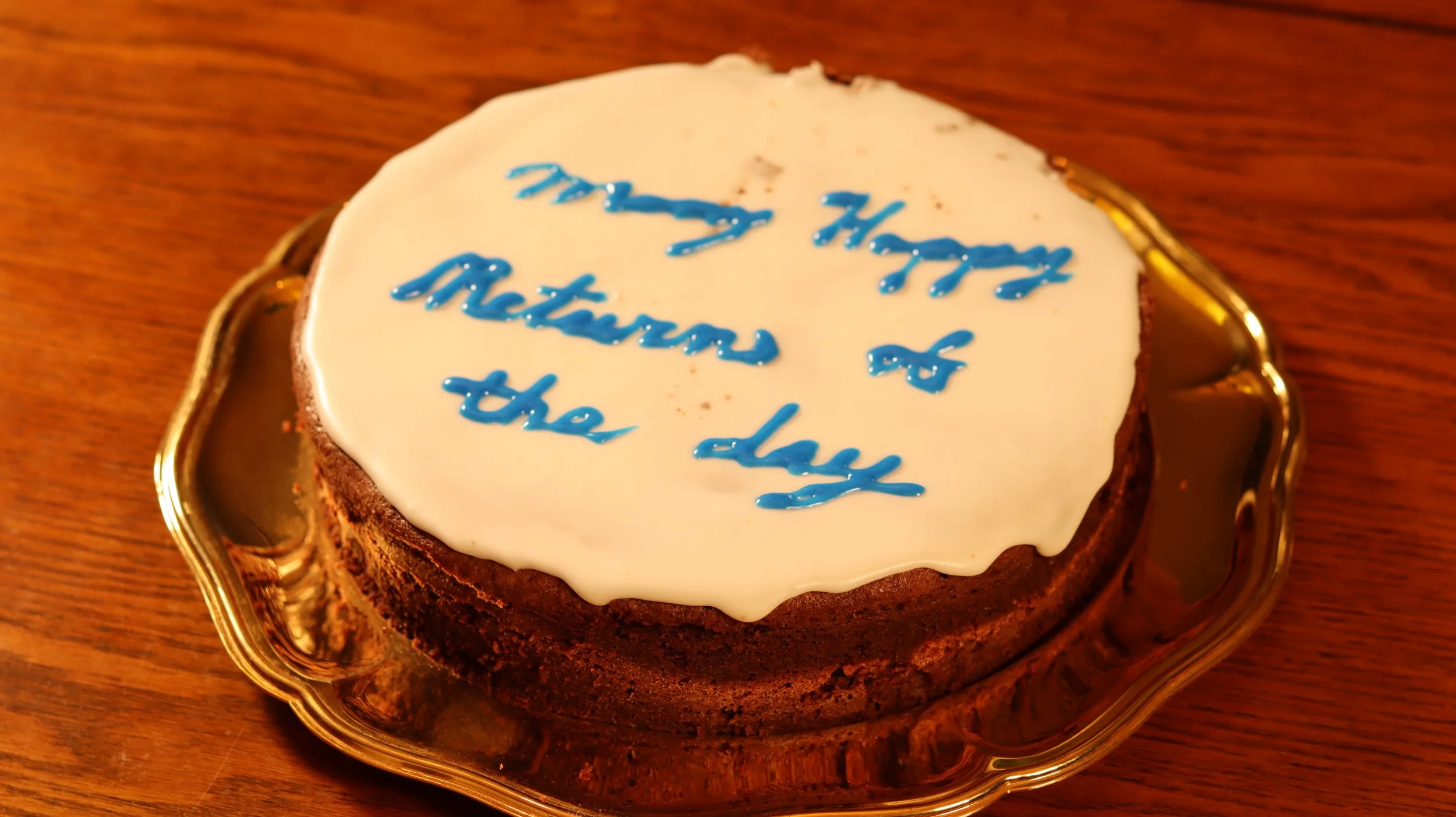 chocolate cake with white icing and "many happy returns of the day" written in blue icing