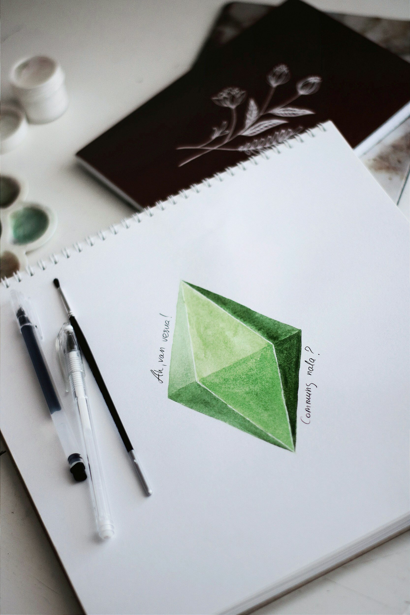 A notebook with a watercolour painting of the plumbob from the Sims with other notebooks and art supplies in the background.