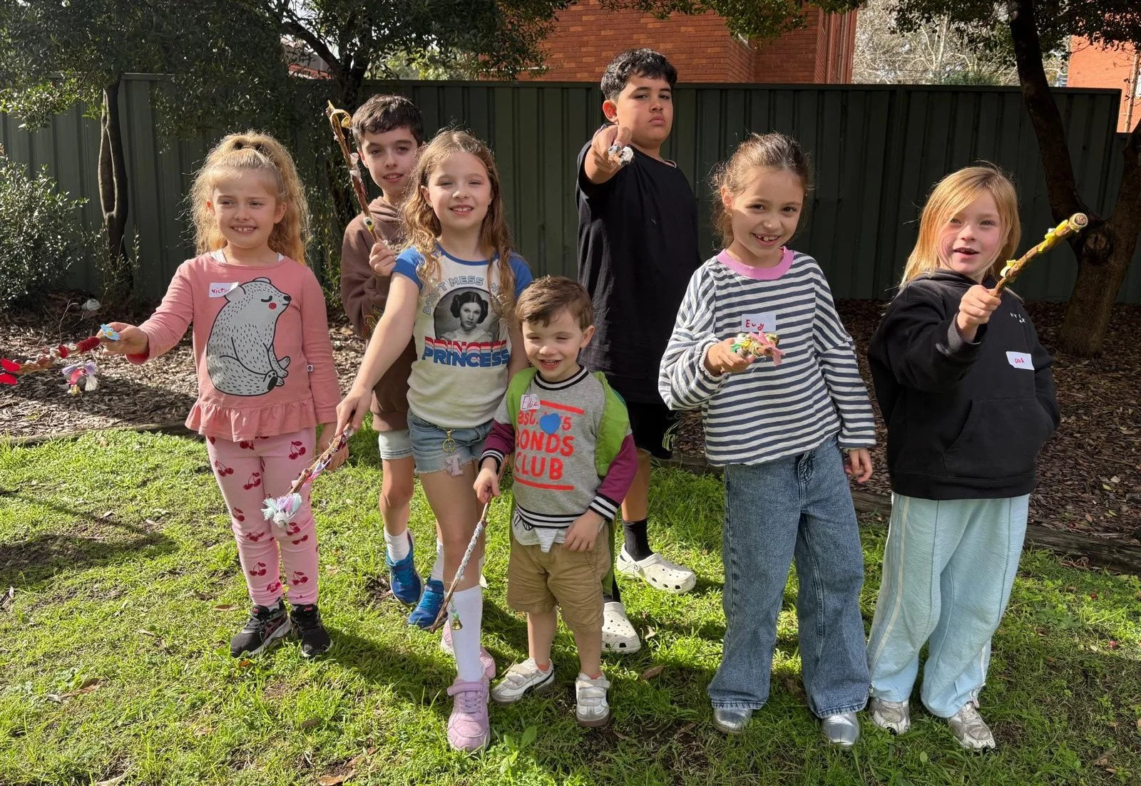 Group of children outdoors holding colorful party blowers, smiling and posing for a photo on a grassy area with trees and a fence in the background.