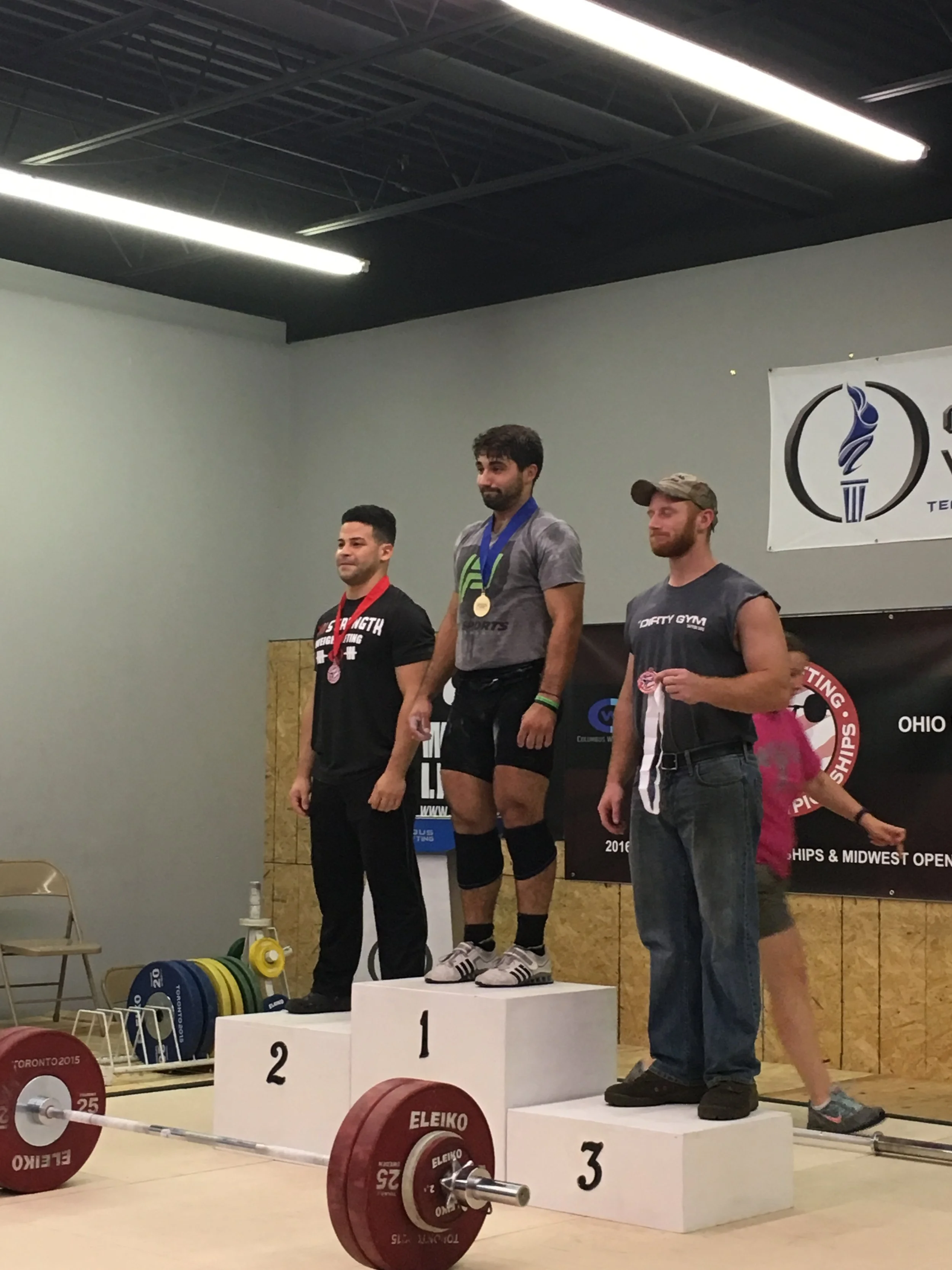A weightlifter on the first-place podium holding a gold medal, flanked by second and third-place winners on podiums. A barbell with weights is in the foreground. The setting is a gym or competition venue with banners and weightlifting equipment visib