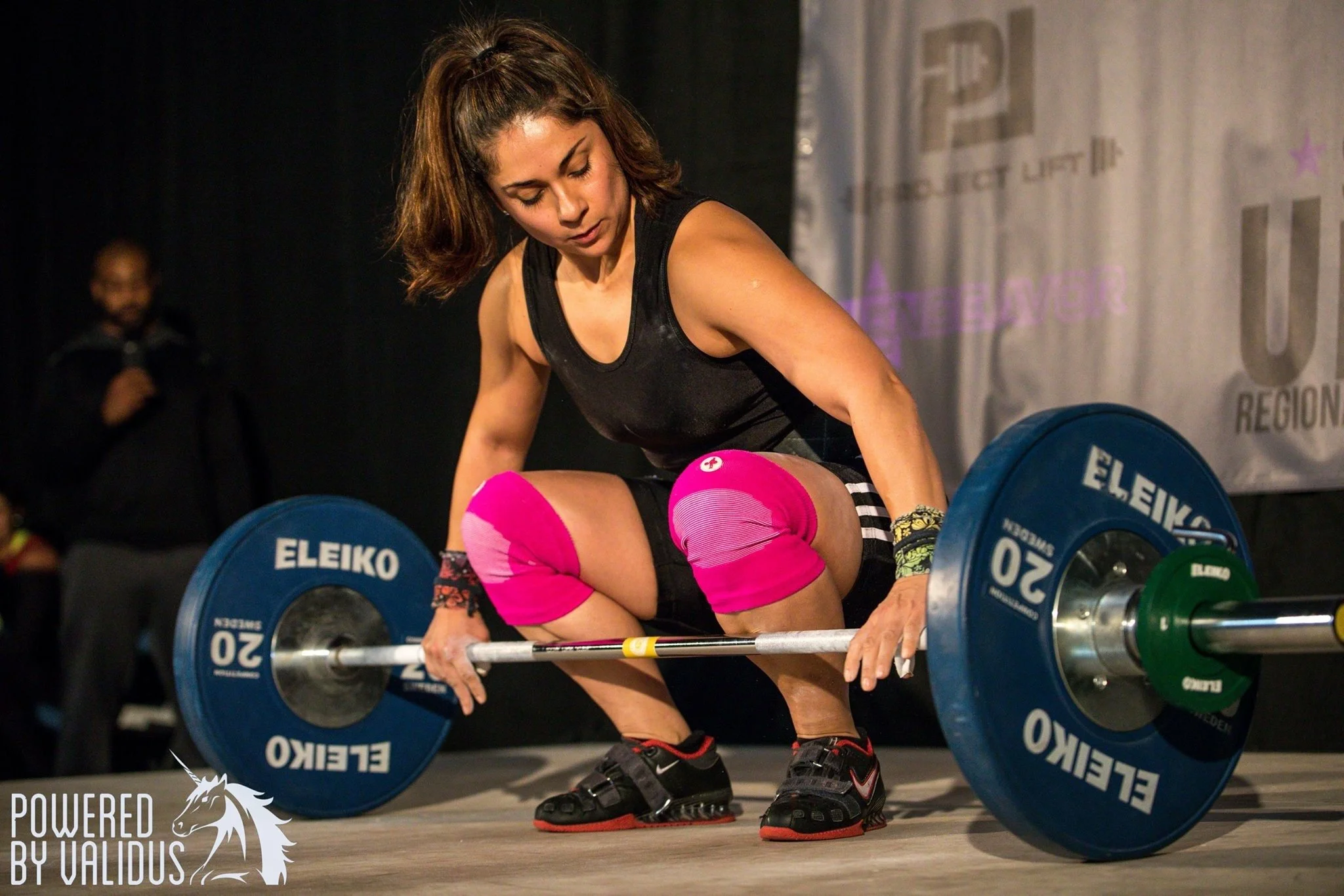 A woman with brown hair in a ponytail lifts a barbell with blue weights at a weightlifting competition, wearing black athletic clothing and pink knee sleeves.