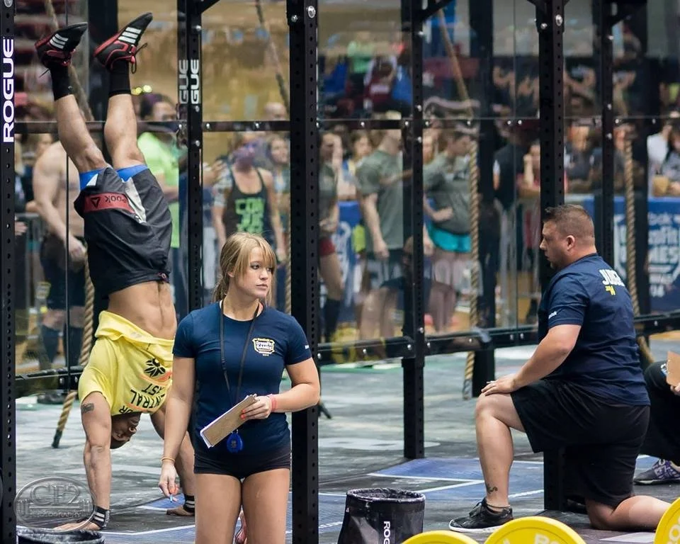 A man doing a handstand against a gym structure during a CrossFit event, with a woman holding a clipboard and a man sitting on a bench nearby, amidst spectators watching in the background.