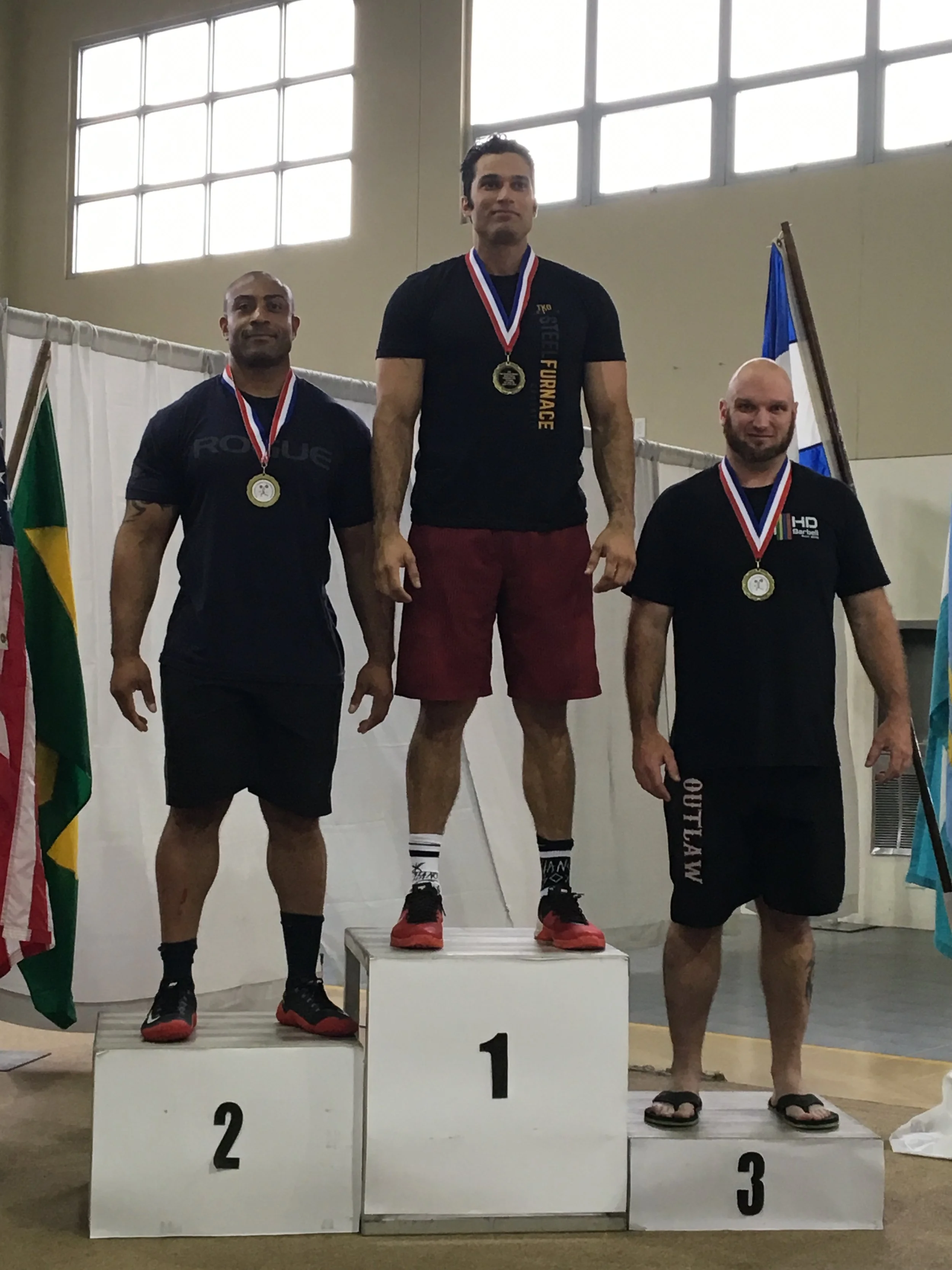 Three men standing on a winners' podium with medals, with a large window behind them and flags on either side. The man in the center is on the top step, wearing a black shirt with yellow text, red shorts, and a medal. The man on the left is on the se