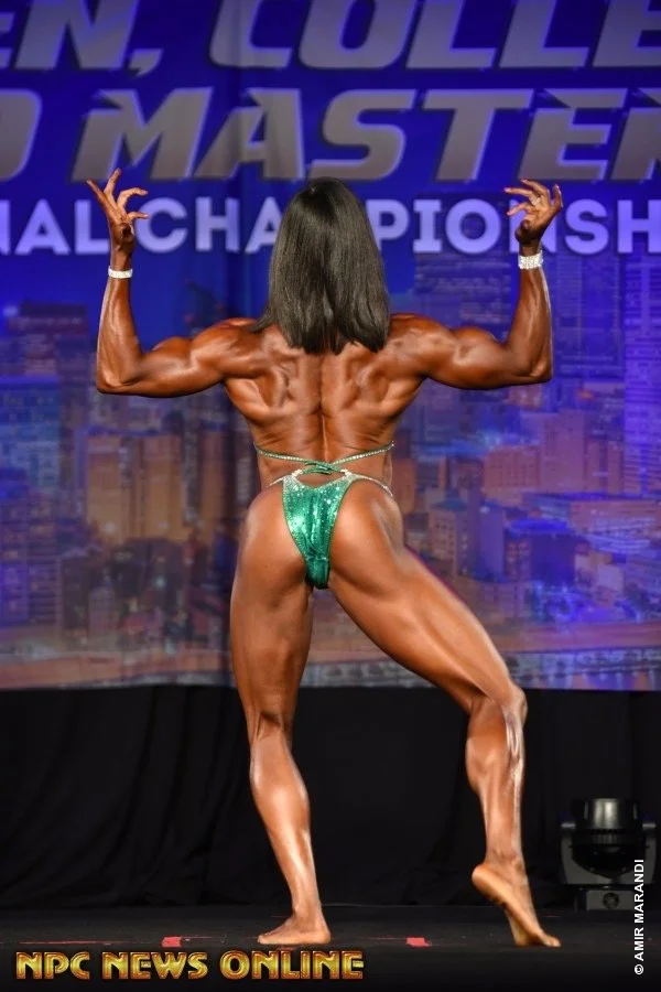 A female bodybuilder flexes her back muscles on stage during a bodybuilding competition, wearing a green sparkly bikini and a silver watch, with a cityscape backdrop and the text 'NPC News Online' at the bottom.