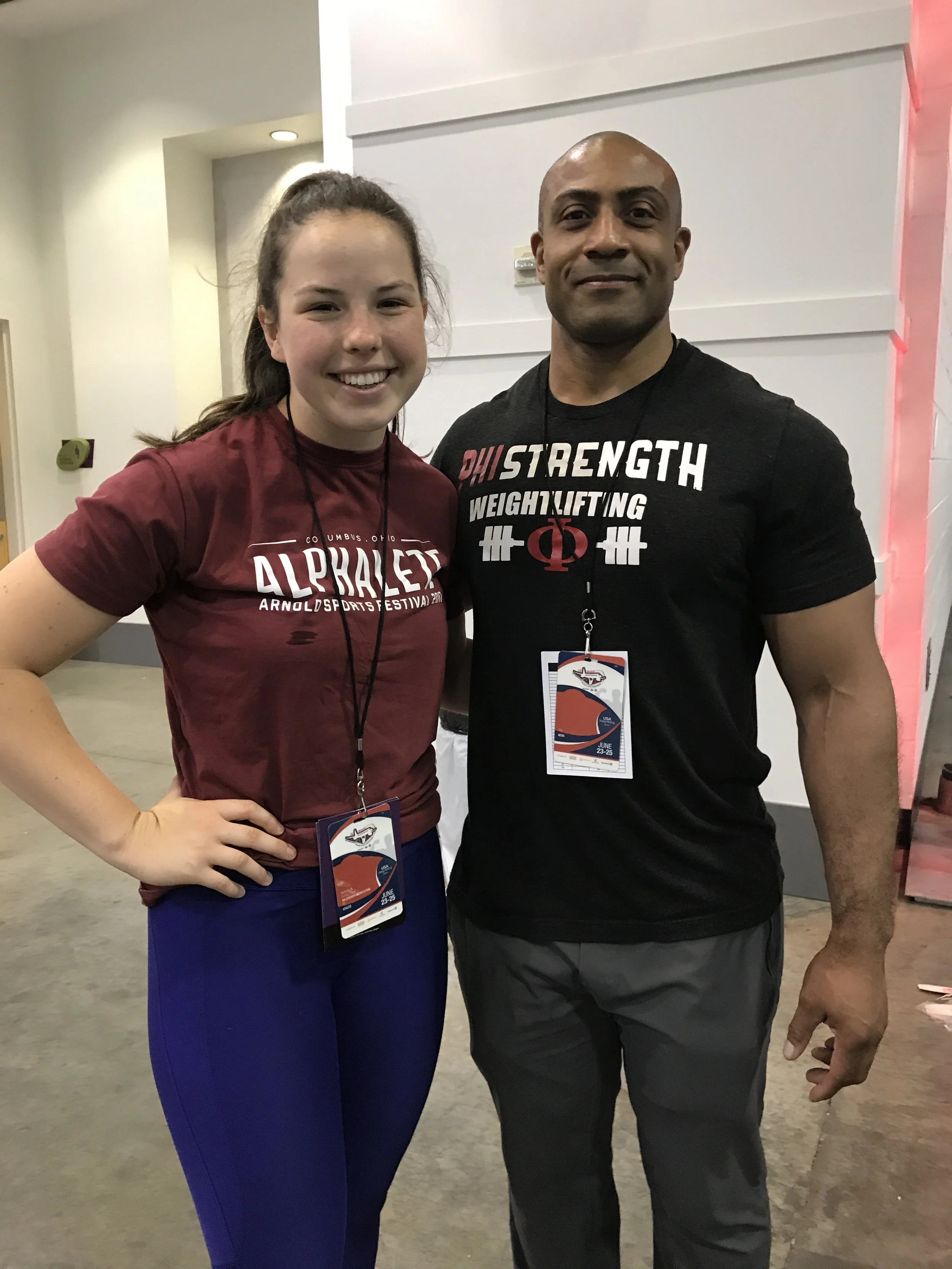 A smiling young woman with long brown hair wearing a maroon athletic shirt and blue leggings stands next to a smiling man with a shaved head wearing a black T-shirt with weightlifting and strength words. Both are wearing event badges hanging around t