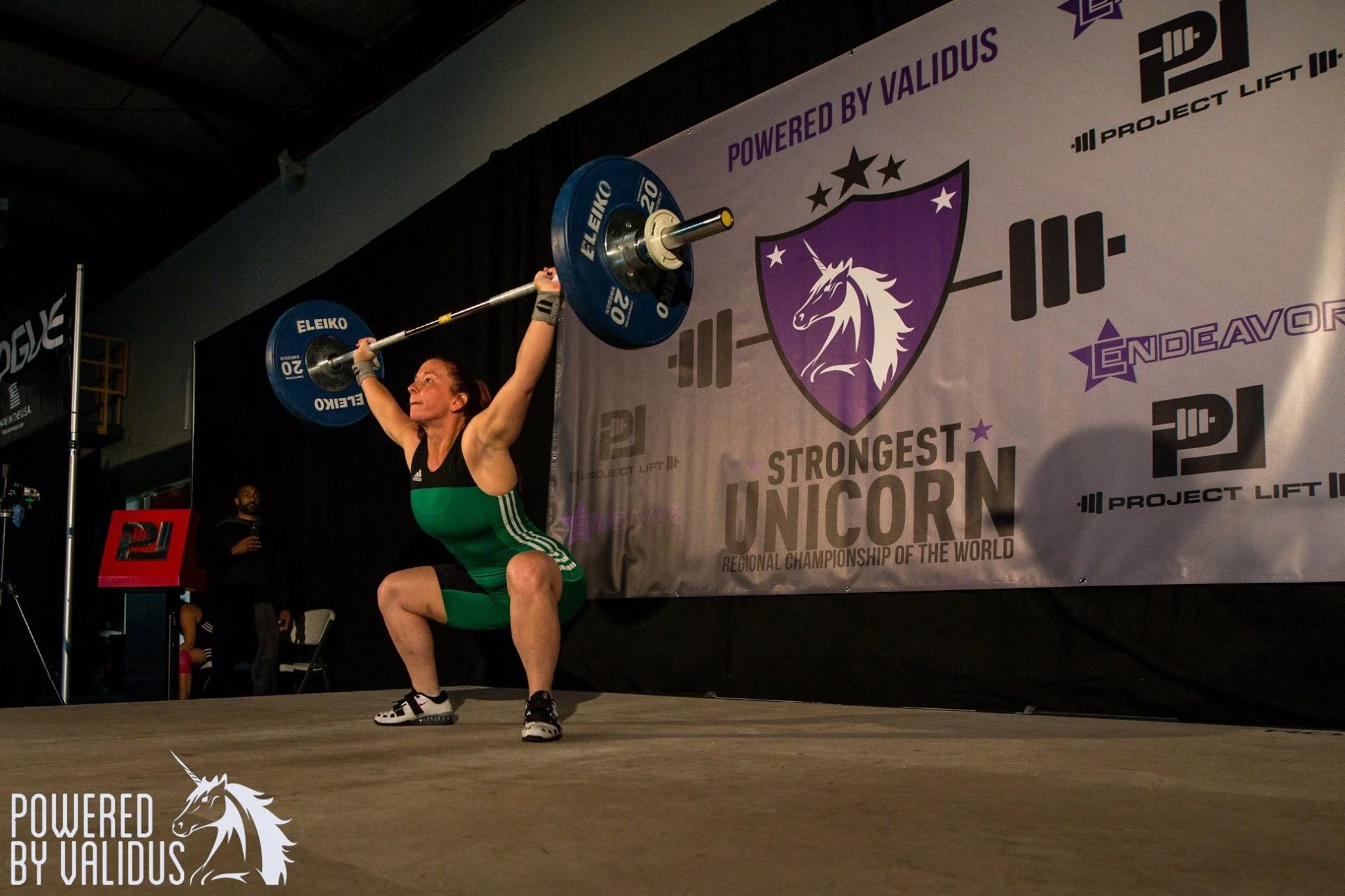 Female weightlifter in black and green attire performing a squat lift with a loaded barbell during a competition, with a banner in the background reading 'Strongest Unicorn' and 'Regional Championship of the World'.