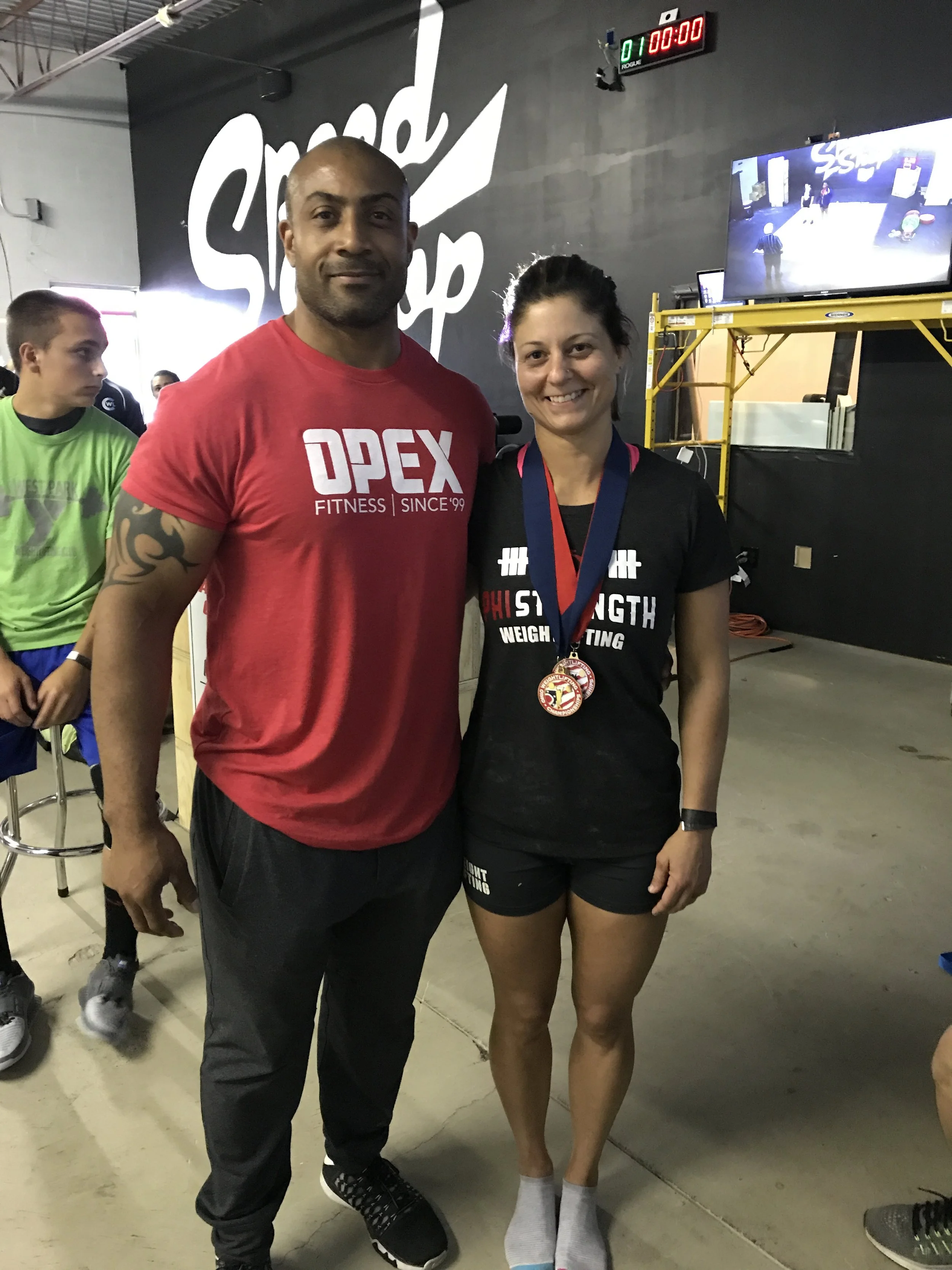 A man and a woman stand together in a gym, smiling. The woman wears a medal around her neck, and the man has his arm around her. The background shows other people, gym equipment, and a large sign that reads 'Sped.'