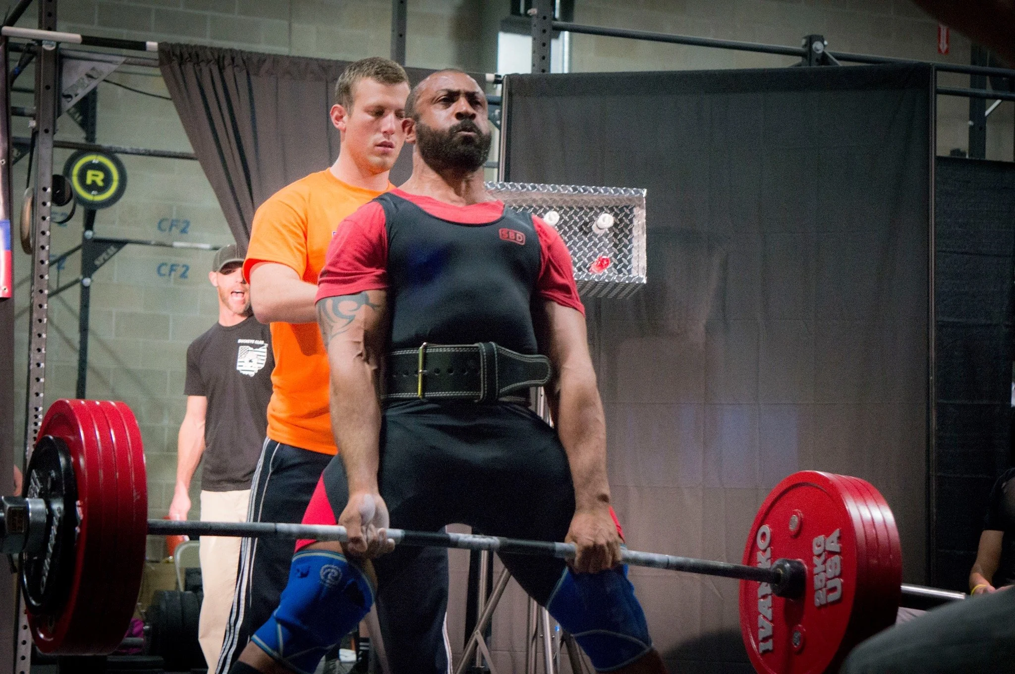 Man performing a deadlift with a loaded barbell at a gym, supervised by a spotter behind him, with other gym members in the background.