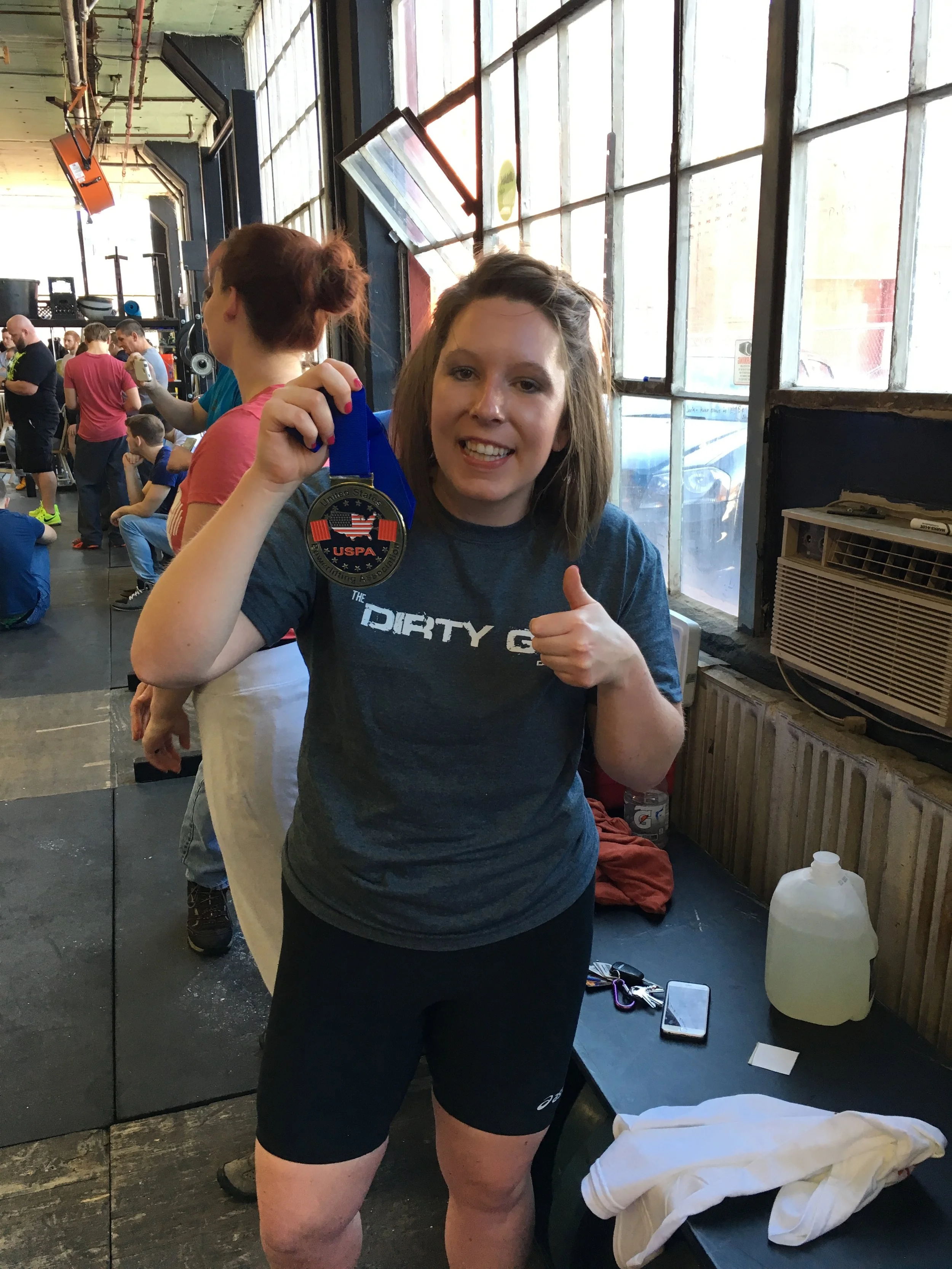 Woman in athletic clothing holding a medal with USPA logo, giving a thumbs-up, inside a gym with other people in the background.