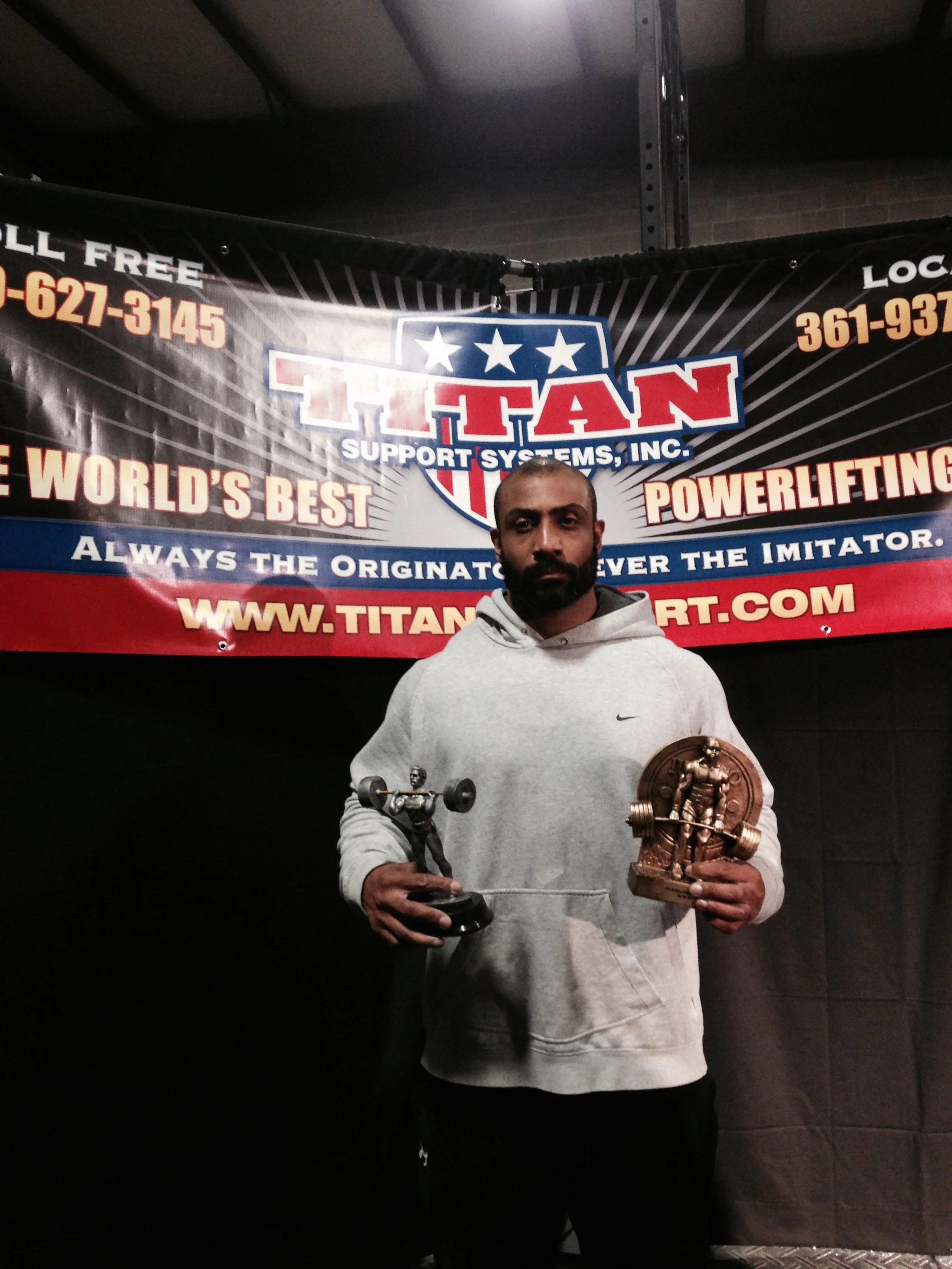 Man standing in front of a banner holding two awards, one in each hand. The banner features the Titan Support Systems, Inc. logo and text about powerlifting.