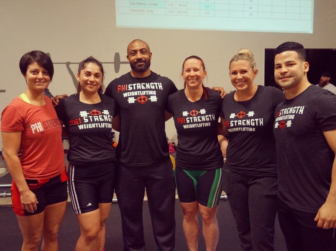 Group of six people in a gym standing side by side, some with arms around each other, wearing workout clothes with 'PH Strength Weightlifting' logo, in front of a wall with a projection screen.