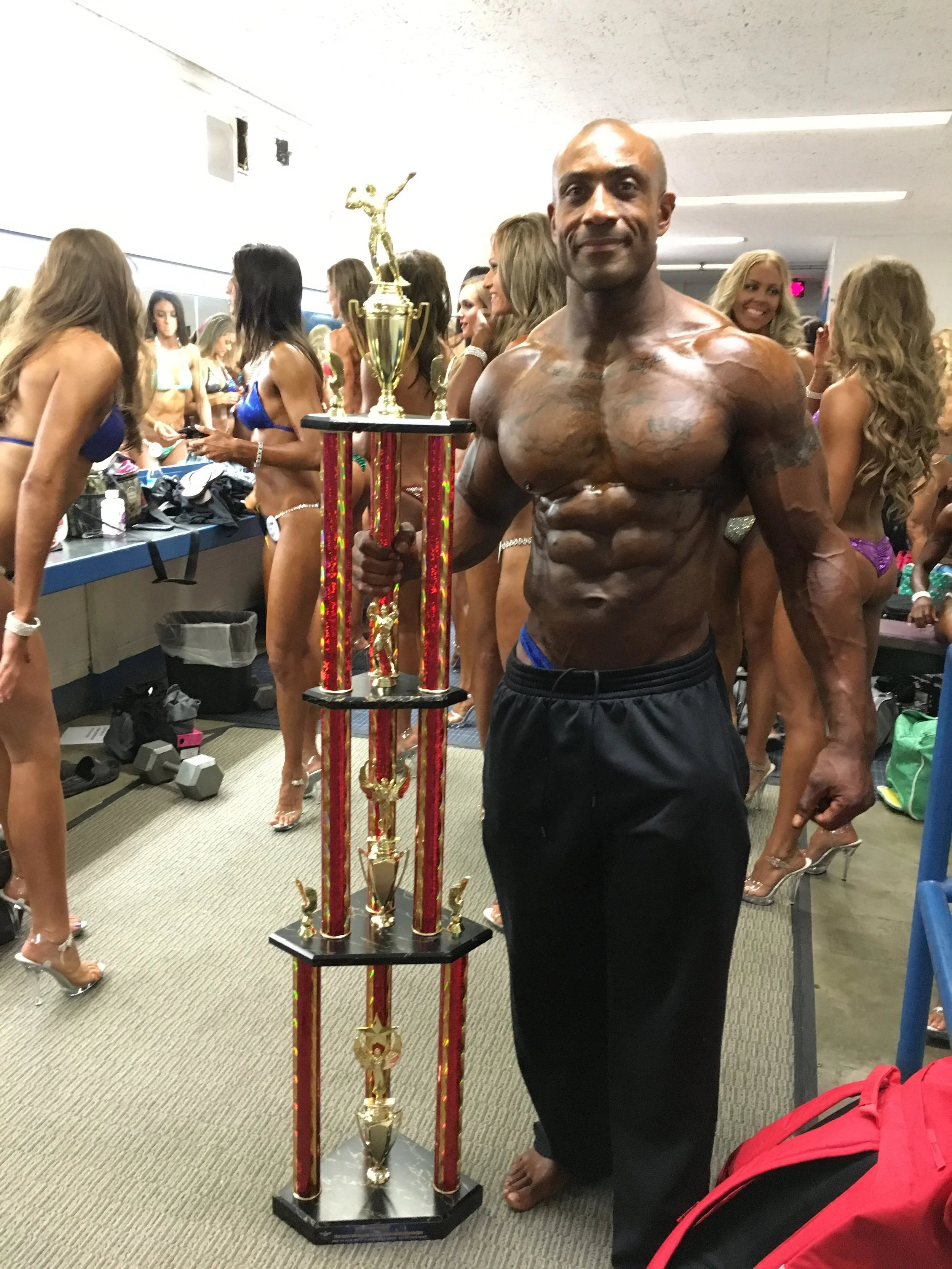 A muscular man with tattoos holding a tall trophy, standing in a backstage area with women in bikinis in the background at a bodybuilding competition.