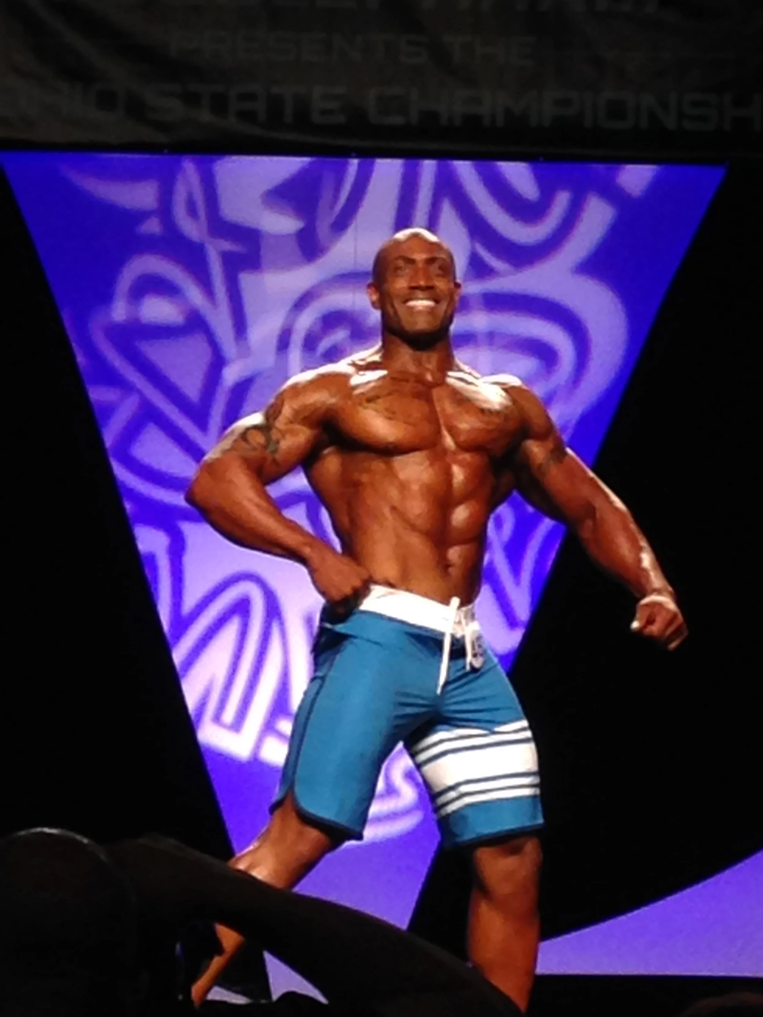 A muscular man on stage at a bodybuilding competition, posing shirtless and wearing blue and white shorts, with a purple and black background.